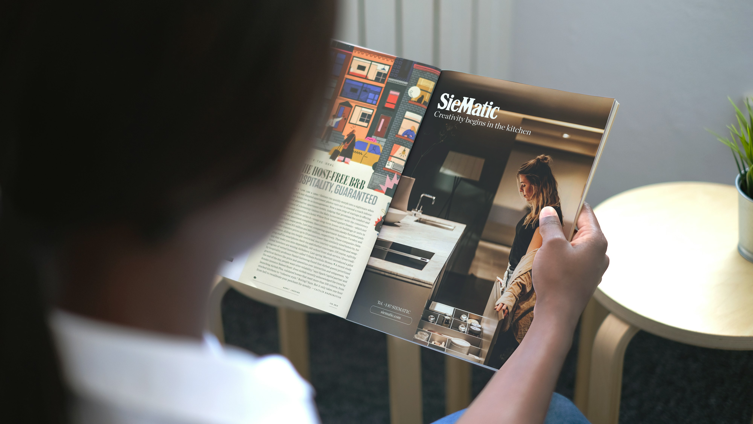 Person reading a magazine with interior design and kitchen images, sitting near a small wooden table and a potted plant.
