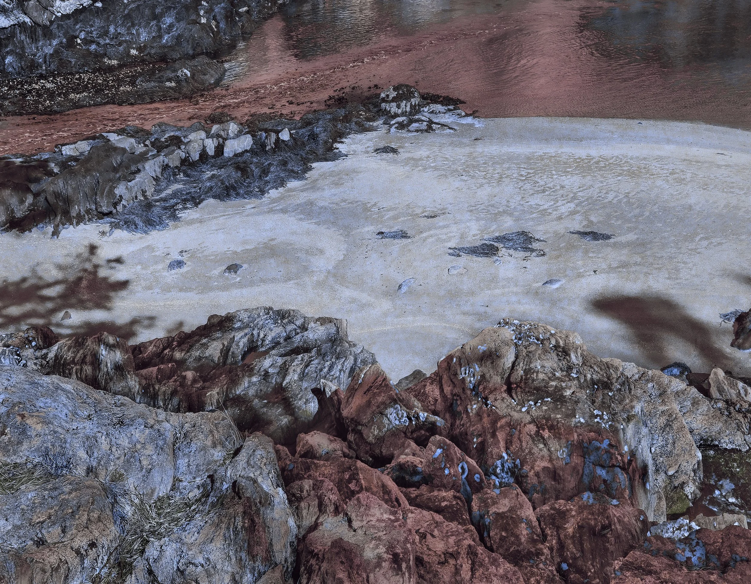 A rocky seashore with brown and gray stones, sand, and water, possibly during sunset or dusk.
