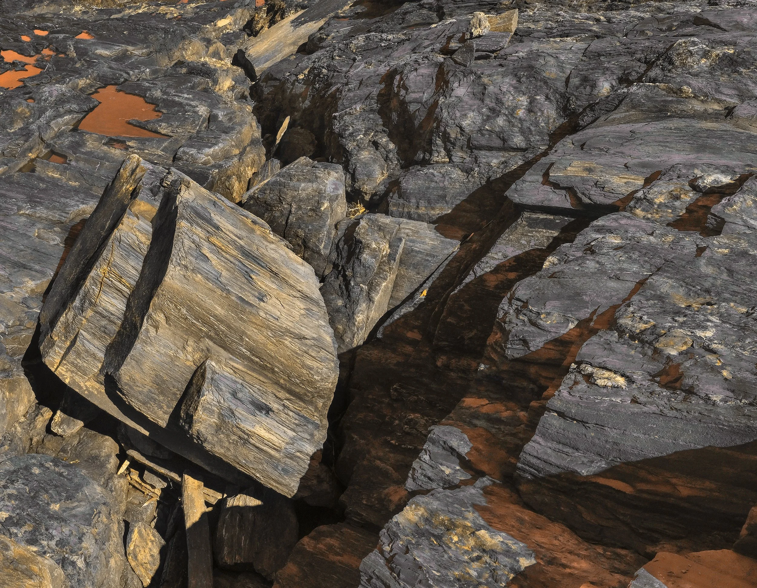Close-up of layered, dark, volcanic rocks with some reddish-brown patches and cracks, illuminated by warm sunlight.