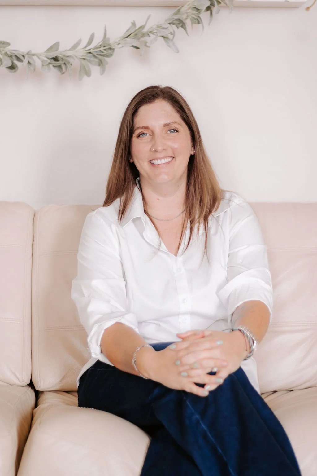 A woman with brown hair, wearing a white shirt and dark pants, sitting on a light-colored sofa with a neutral background and decorative greenery hanging on the wall behind her.