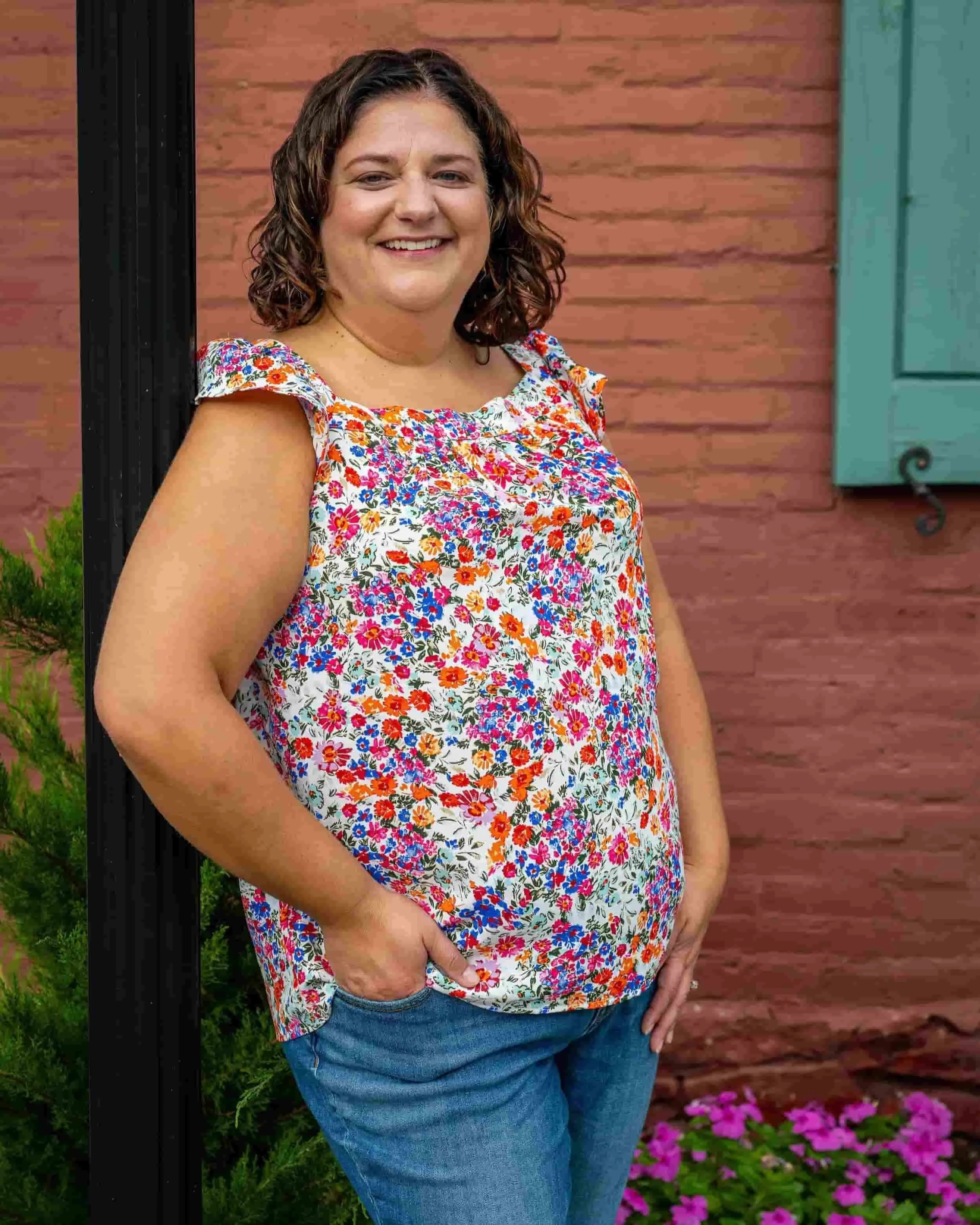 Woman with curly brown hair, wearing a floral top and jeans, smiles while standing outside near a brick wall and green shutter.