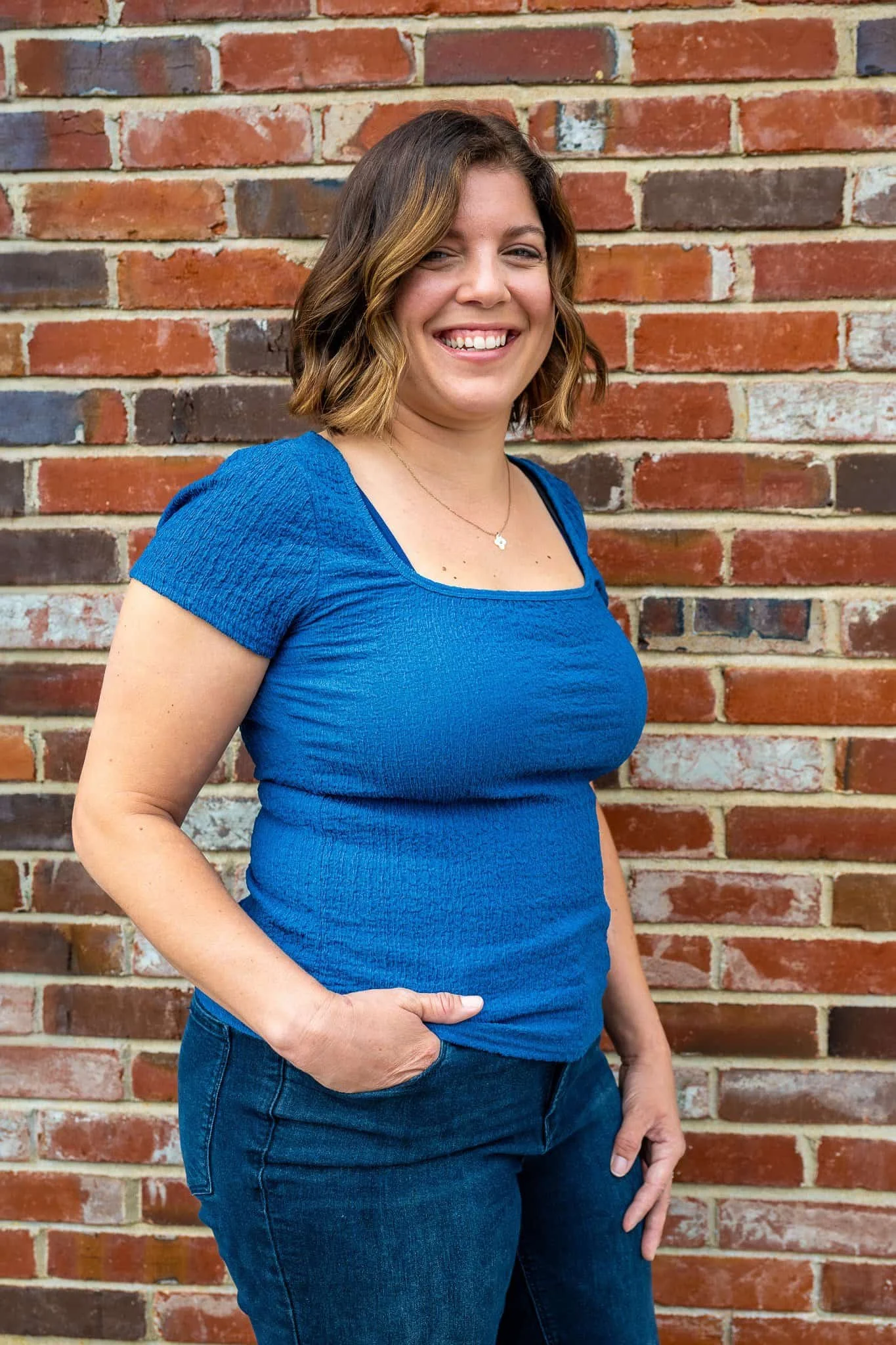 A woman with shoulder-length wavy brown hair, wearing a bright blue peasant-style top and dark jeans, standing against a brick wall, smiling and looking at the camera.