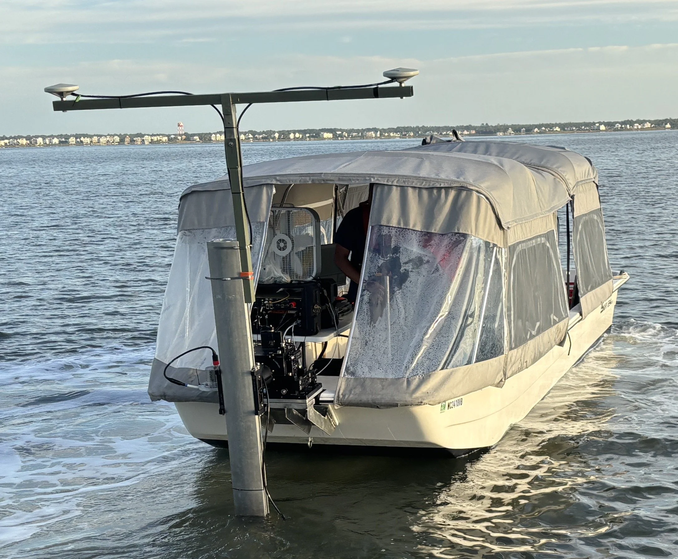 A boat with a shelter on the water, equipped with electronic gear and an antenna, near a shoreline with buildings and cloudy sky in the background.