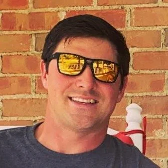 A man with dark hair wearing reflective sunglasses and a dark gray shirt, smiling outdoors in front of a brick wall.