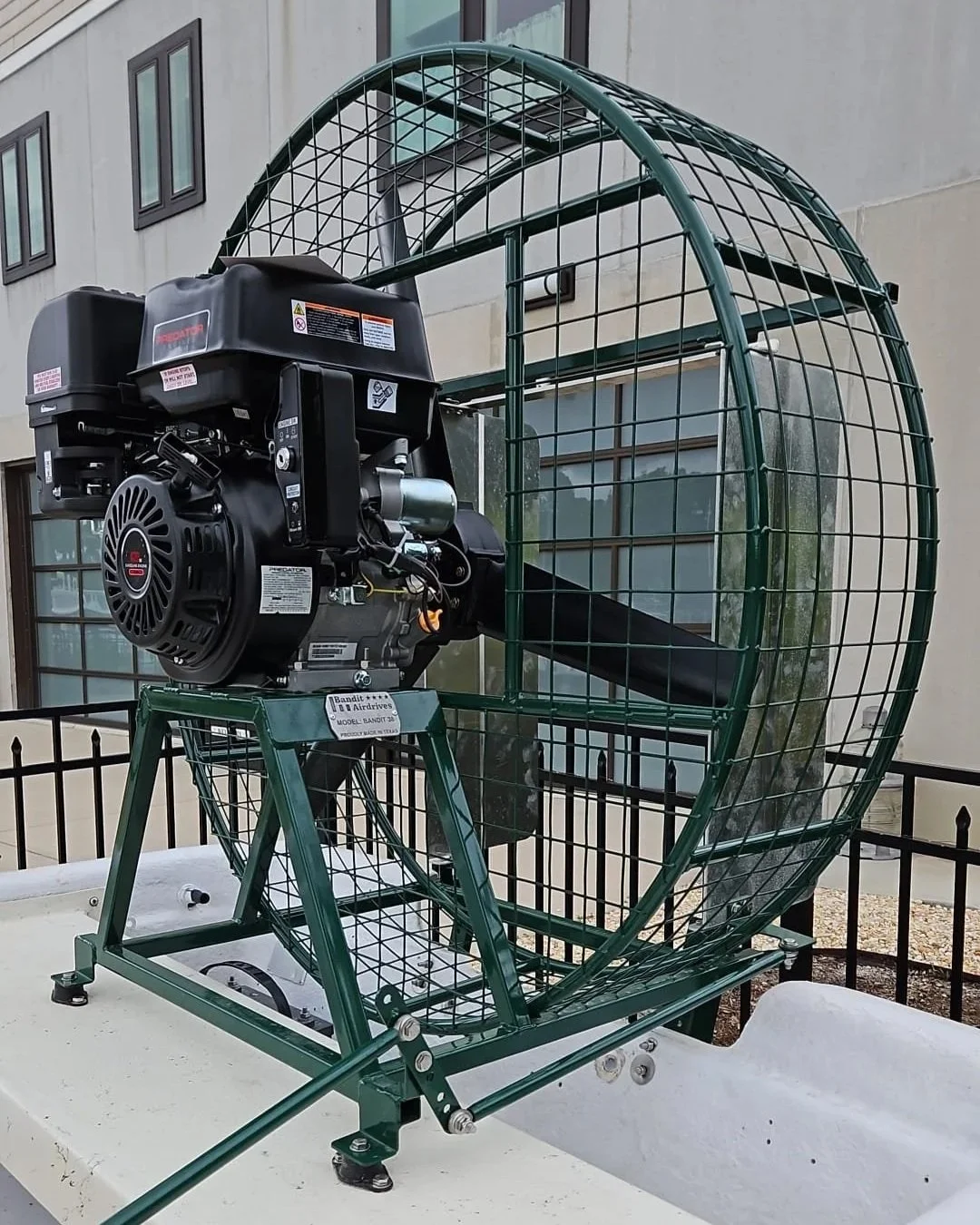 A large portable fan with a protective metal mesh cover and a pull-start engine, mounted on a green metal frame, placed outdoors in front of a building.