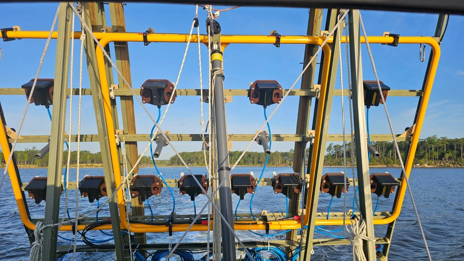 A large frame with multiple devices and wires set up over water, likely part of a scientific or technological installation, with trees along the shoreline in the background.