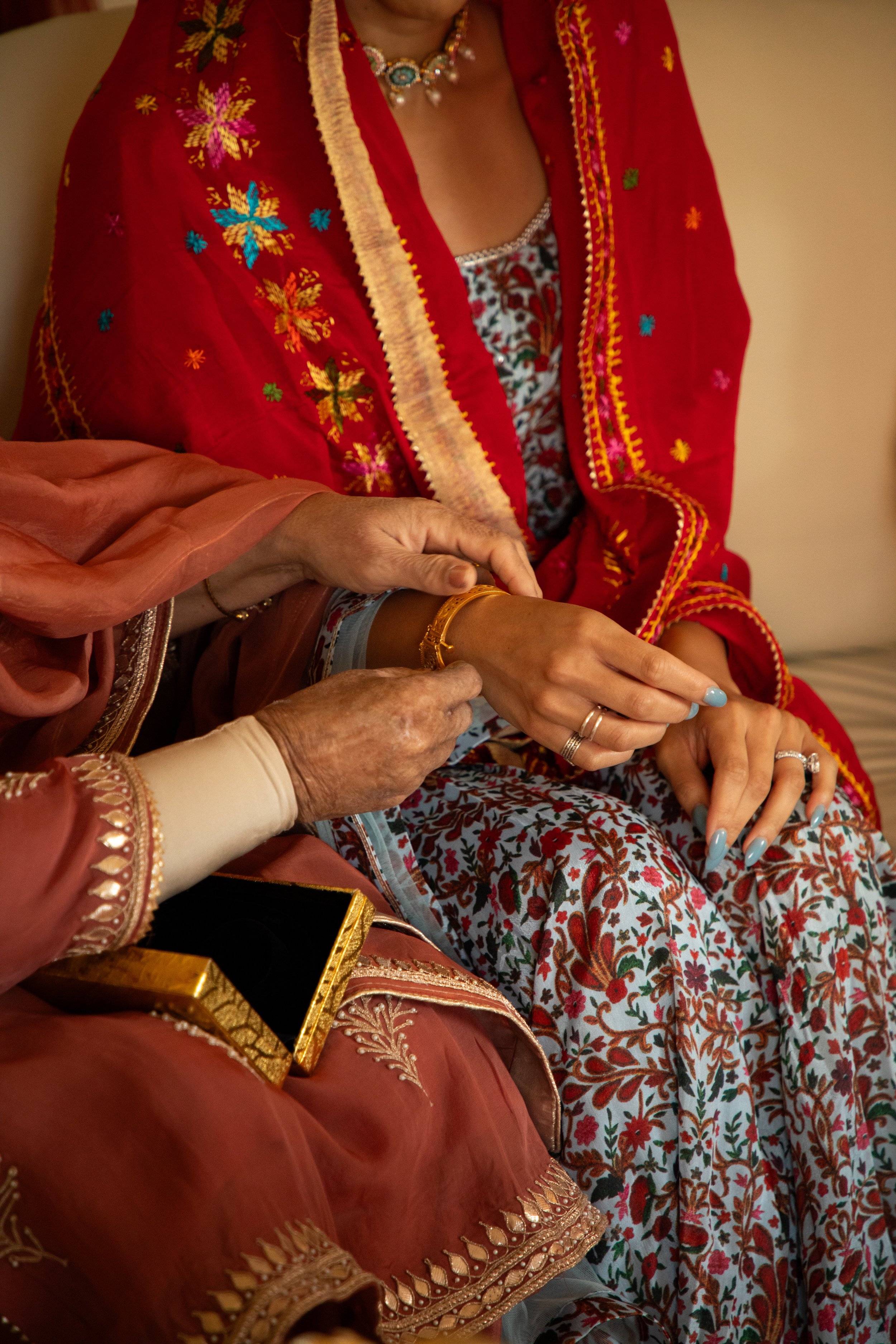 Photo of a woman at a Traditional Indian Engagement Ceremony with traditional Indian robes in shades of red and blue. Her Mother is securing a red string bracelet on her wrist.