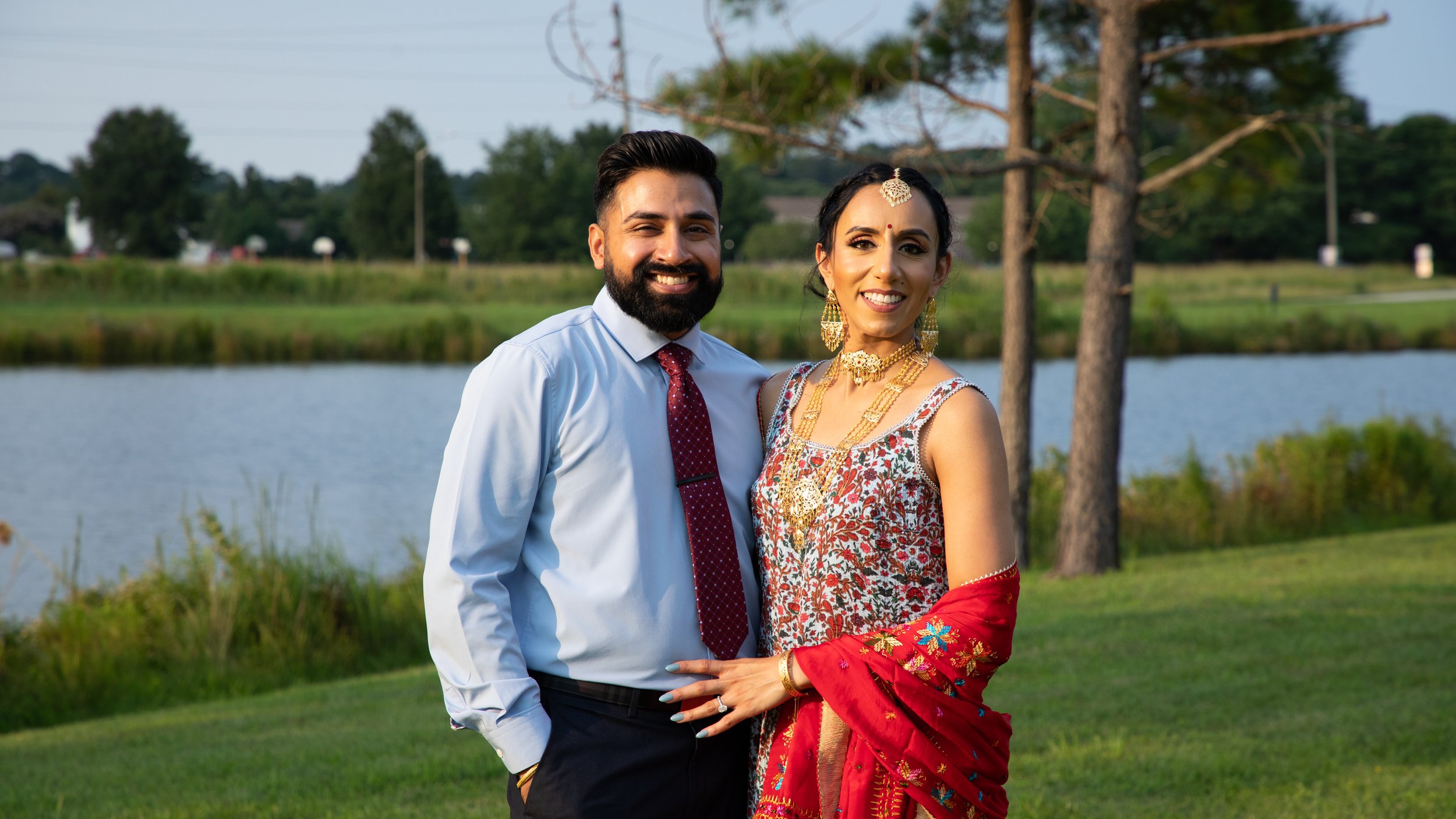 Photo of a newly engaged couple smiling together as they celebrate with a a Traditional Indian Engagement Ceremony. The woman is wearing beautiful gold jewelry and adornments, with traditional Indian robes in shades of red and blue.