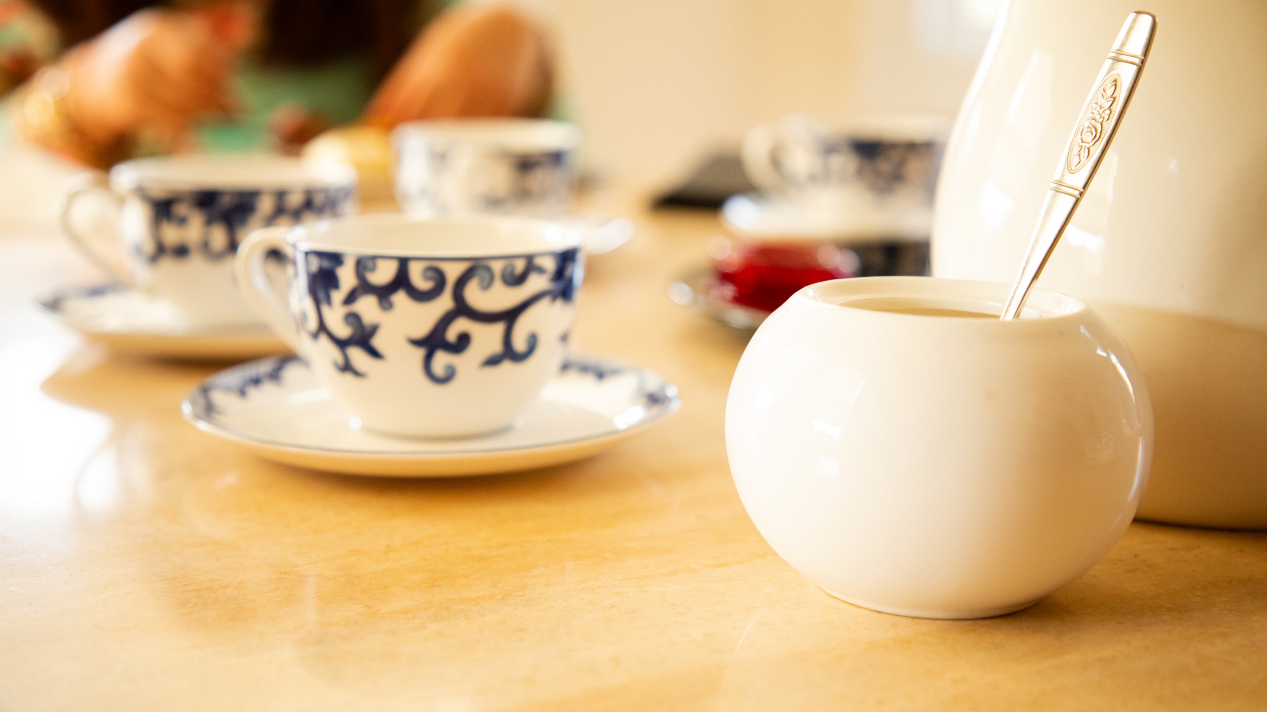 Close up photo of beautiful, ornamentally painted tea cups next to a sugar jar with a spoon coming out of it.