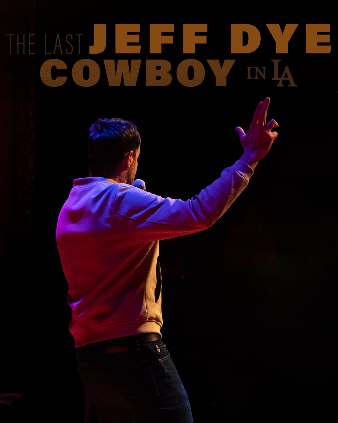 Photo of comedian Jeff Dye performing a live event during his tour, The Last Cowboy in L.A. He has his right arm in the air with his fingers making a finger gun as he tells a joke.