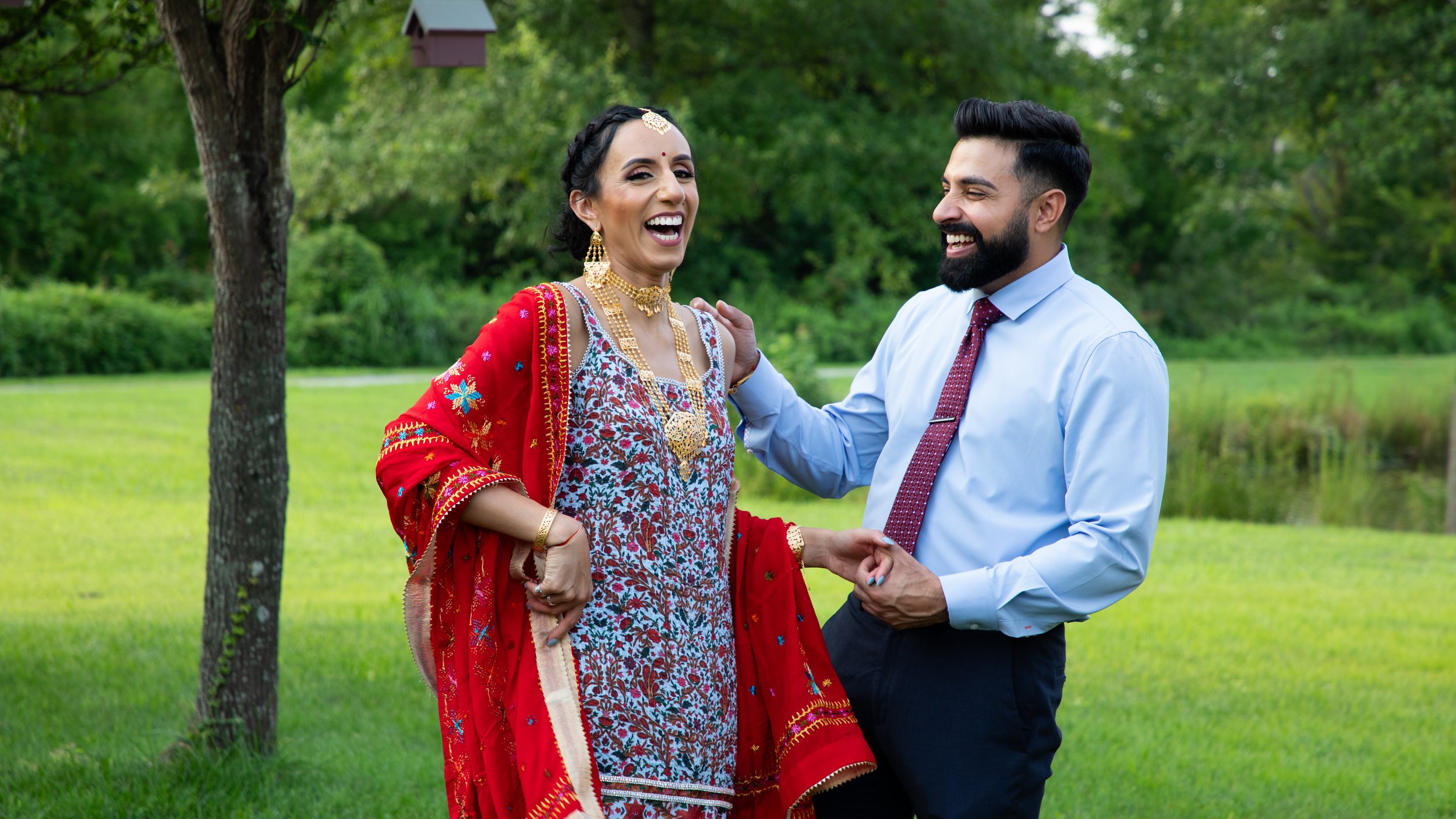 Photo of a newly engaged couple laughing together as they celebrate with a a Traditional Indian Engagement Ceremony. The woman is wearing beautiful gold jewelry and adornments, with traditional Indian robes in shades of red and blue.
