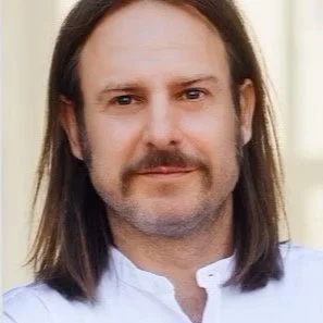 A man with shoulder-length brown hair and a mustache, wearing a white shirt, standing indoors.