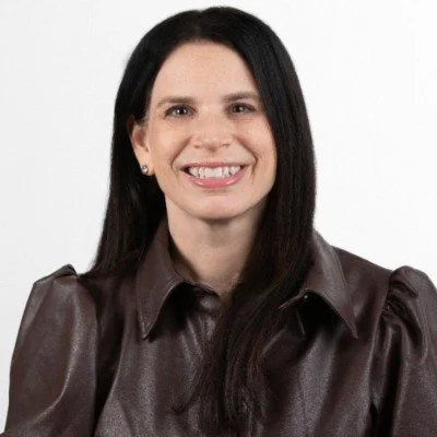 A woman with long dark hair smiling at the camera, wearing a brown leather shirt, against a plain white background.