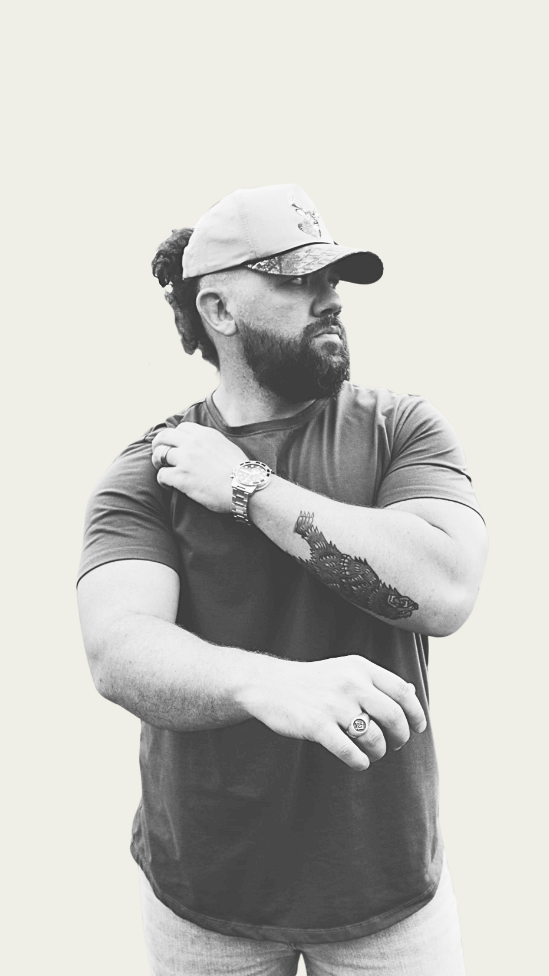 Black and white photo of a man with a beard and dreadlocks, wearing a baseball cap, a T-shirt, and a watch. He is looking to the side with his arms crossed.