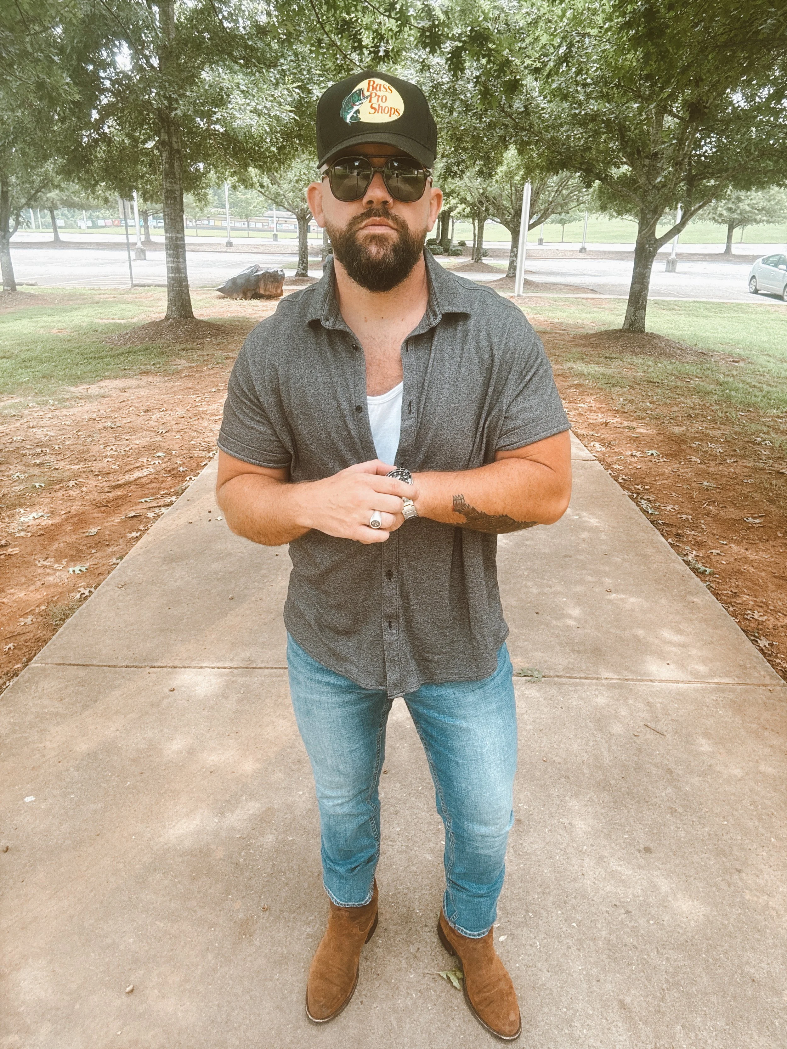 A man standing outdoors on a concrete path, wearing sunglasses, a black Bass Pro Shops cap, a gray short-sleeve button-up shirt over a white T-shirt, blue jeans, and brown boots, with trees and cars in the background.