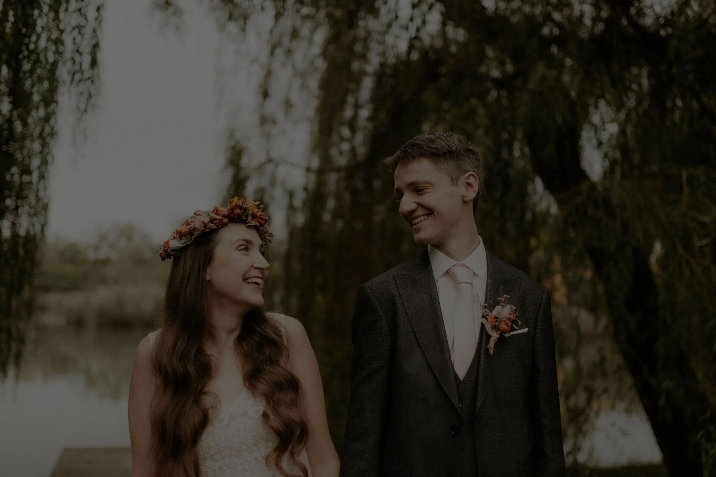 A smiling couple at their wedding outdoors, with a woman wearing a floral crown and a man in a suit with a boutonniere, standing near a body of water surrounded by trees.