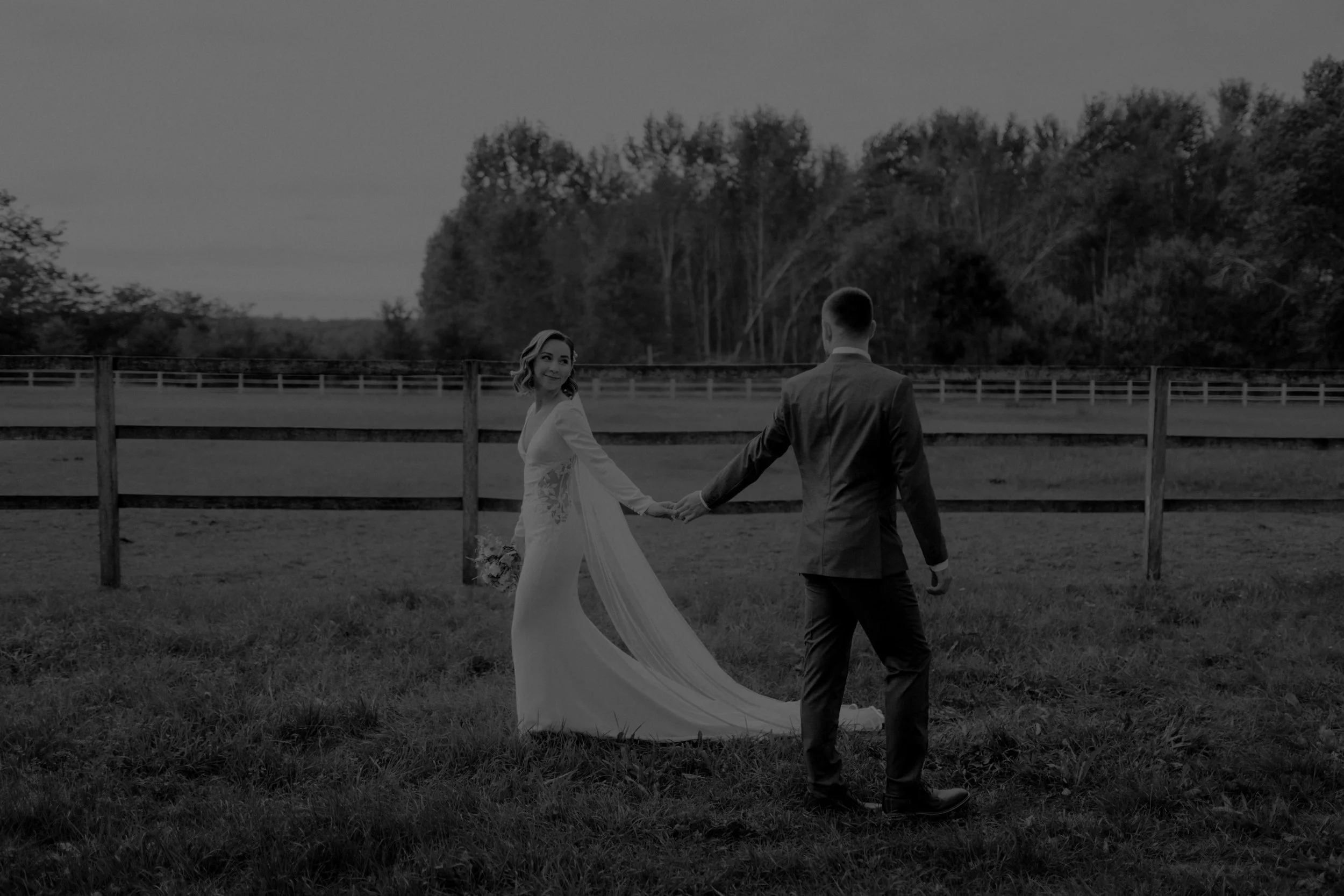 A couple holding hands in a field, the woman wearing a white wedding dress and the man in a suit, with a wooden fence and trees in the background.