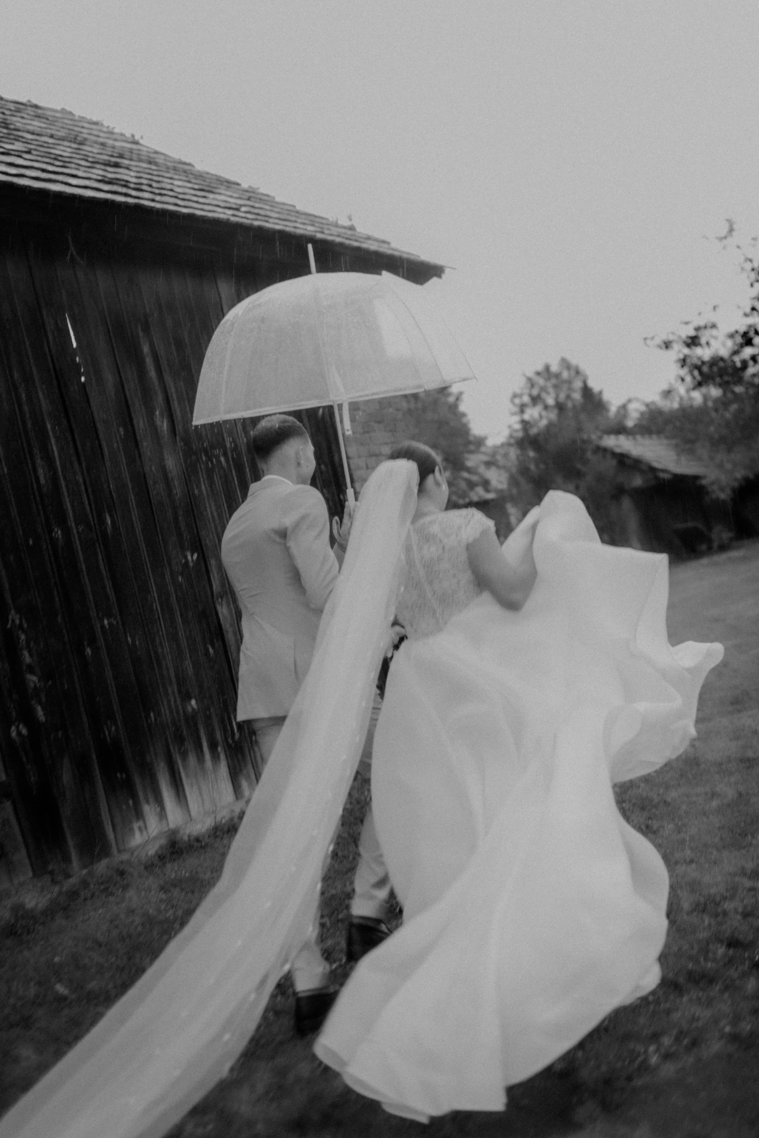 Black and white photo of a bride and groom walking in the rain, holding an umbrella, near a wooden building, with trees in the background.