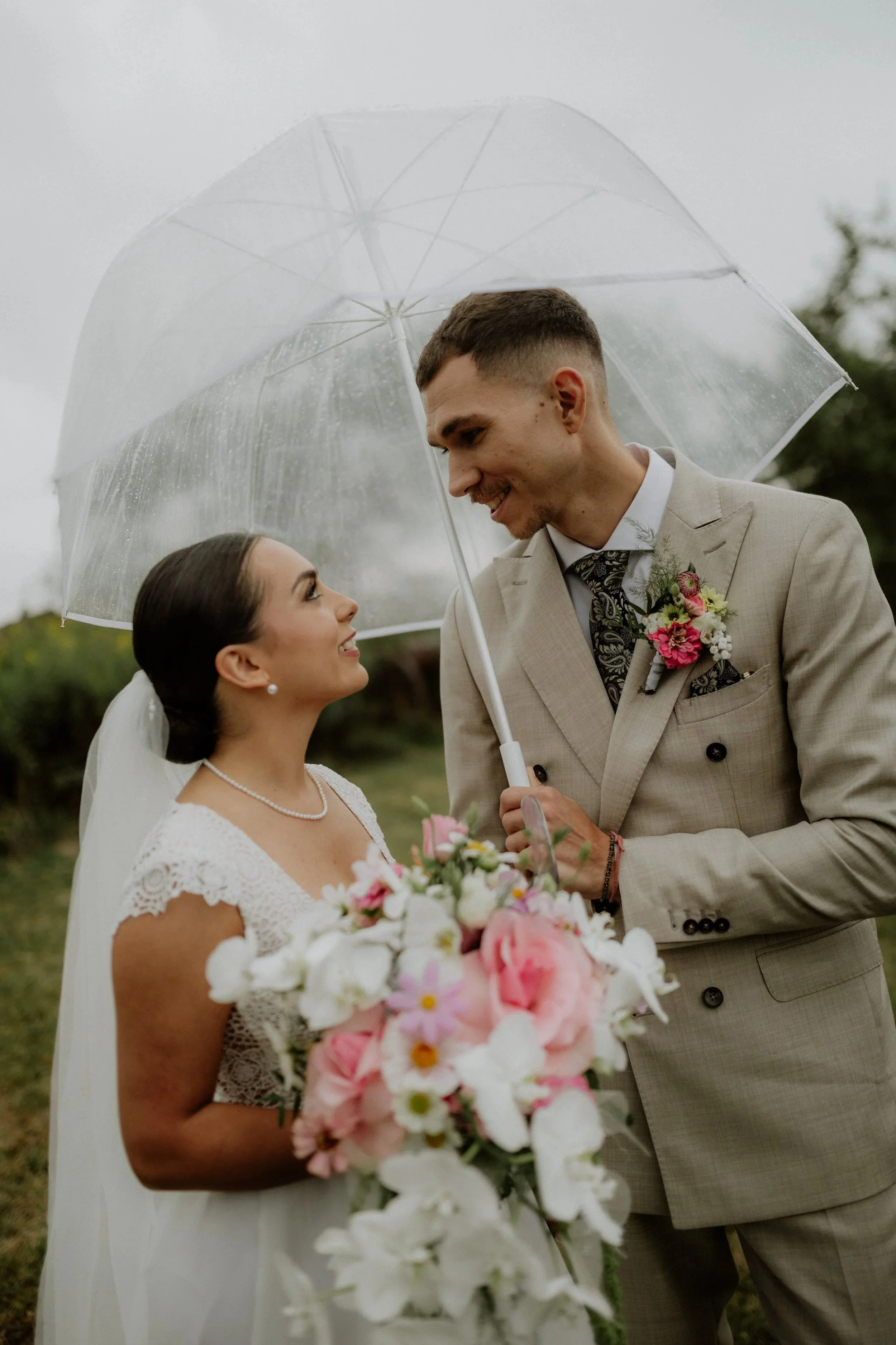 Menyasszony és vőlegény az esküvőjük napján, szabadtéren állva egy átlátszó esernyő alatt, esős időben, a menyasszony kezében rózsaszín és fehér virágokból álló csokorral.