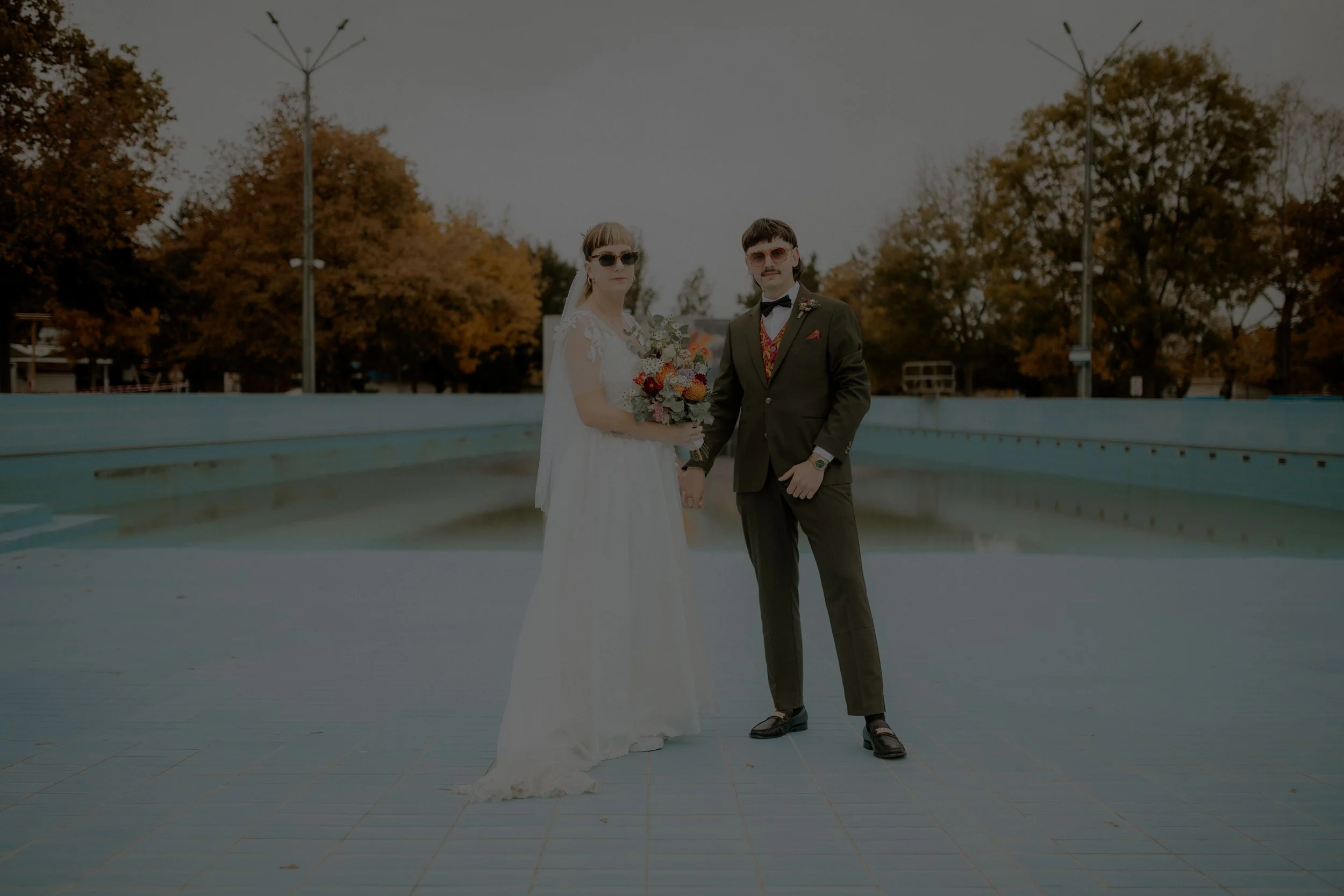 A bride and groom standing in an empty outdoor court with autumn trees in the background, holding hands and facing the camera.