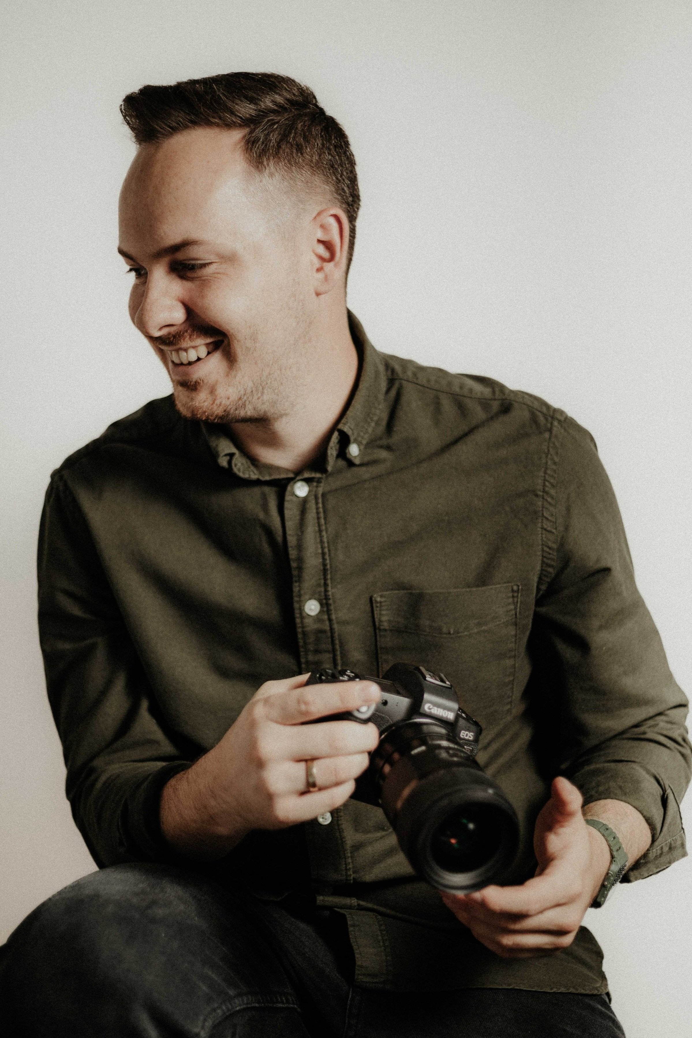 A man in a dark green shirt holding a Canon camera, smiling, looking down.