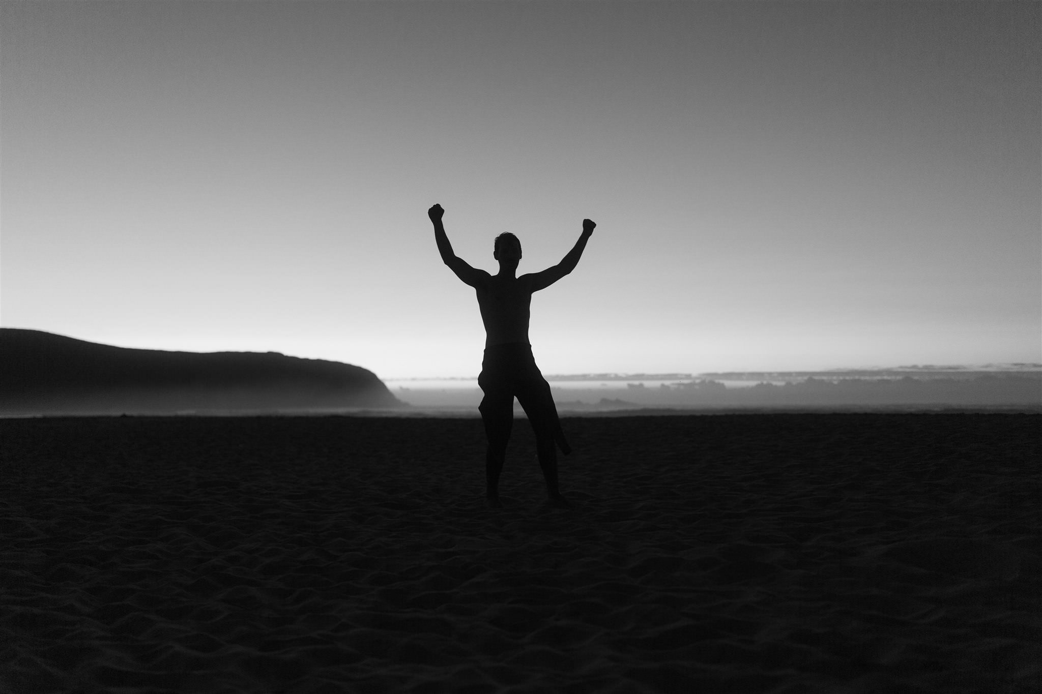 Silhouette of a person with raised arms standing on a beach during sunset or sunrise.