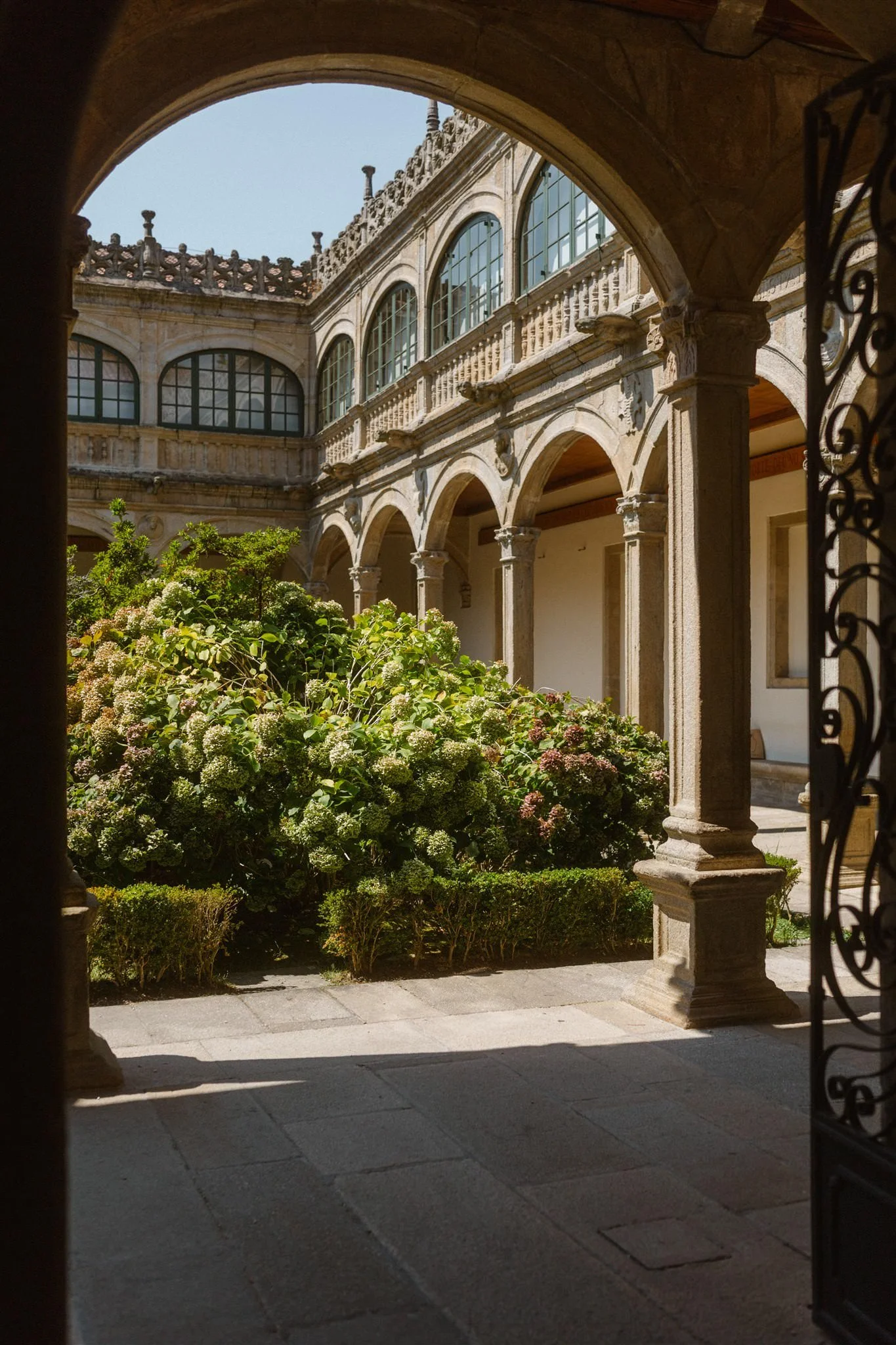 View of a courtyard through an arched entrance, with stone columns, a garden with bushes, and a two-story building with large windows and decorative stonework.