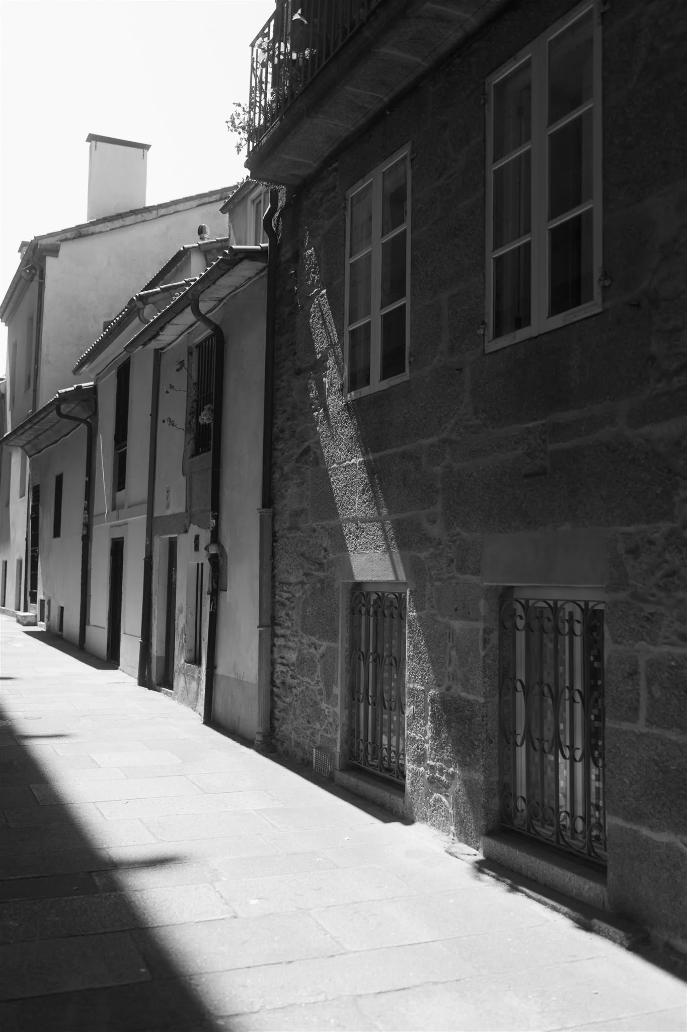 Black and white photo of a narrow street with old buildings, some windows have metal security bars, and the sun casting shadows on the sidewalk.