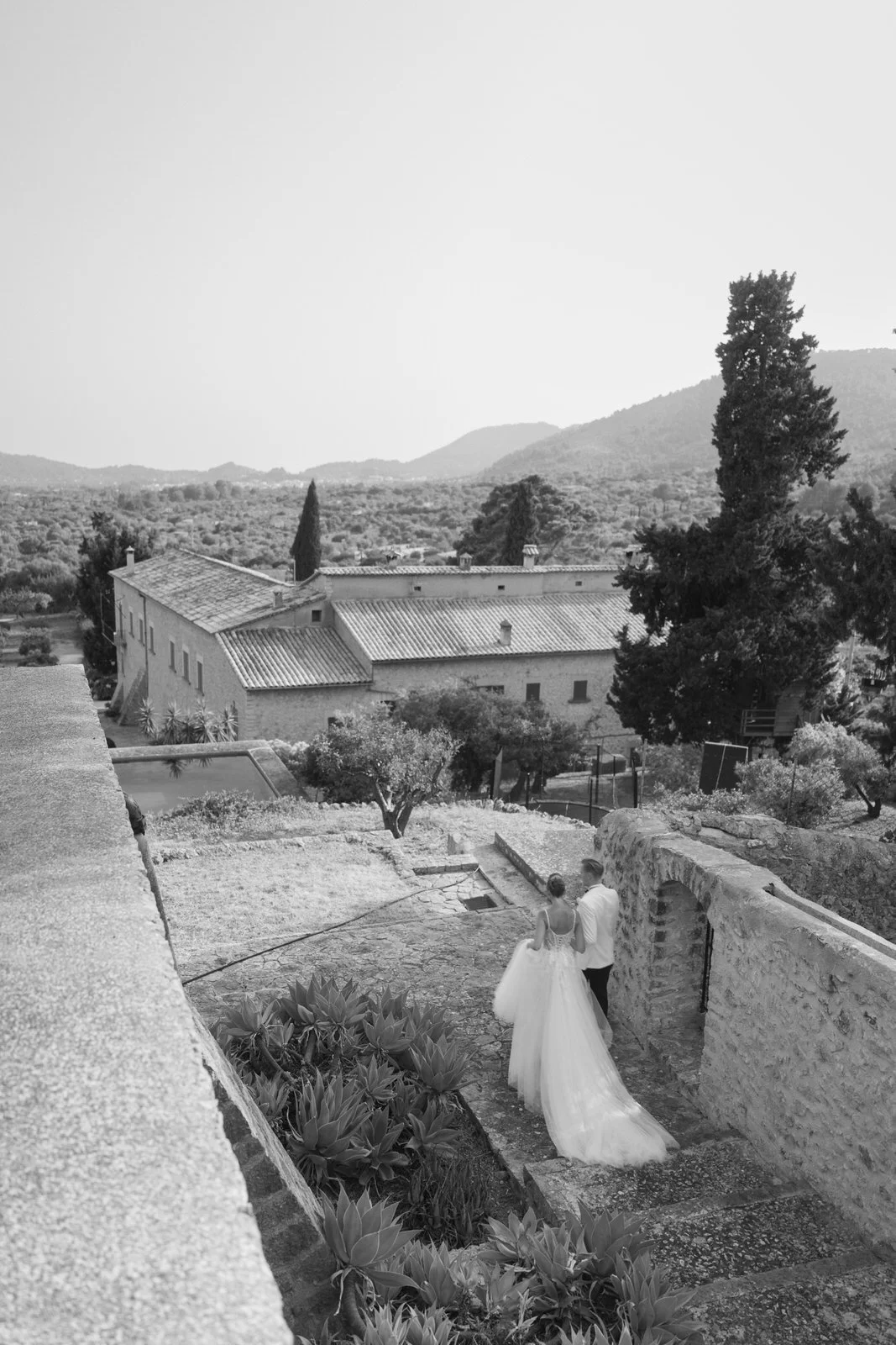 Black and white photo of a bride and groom walking outdoors on a stone pathway near a stone wall in a rural setting with trees, houses, and mountains in the background.