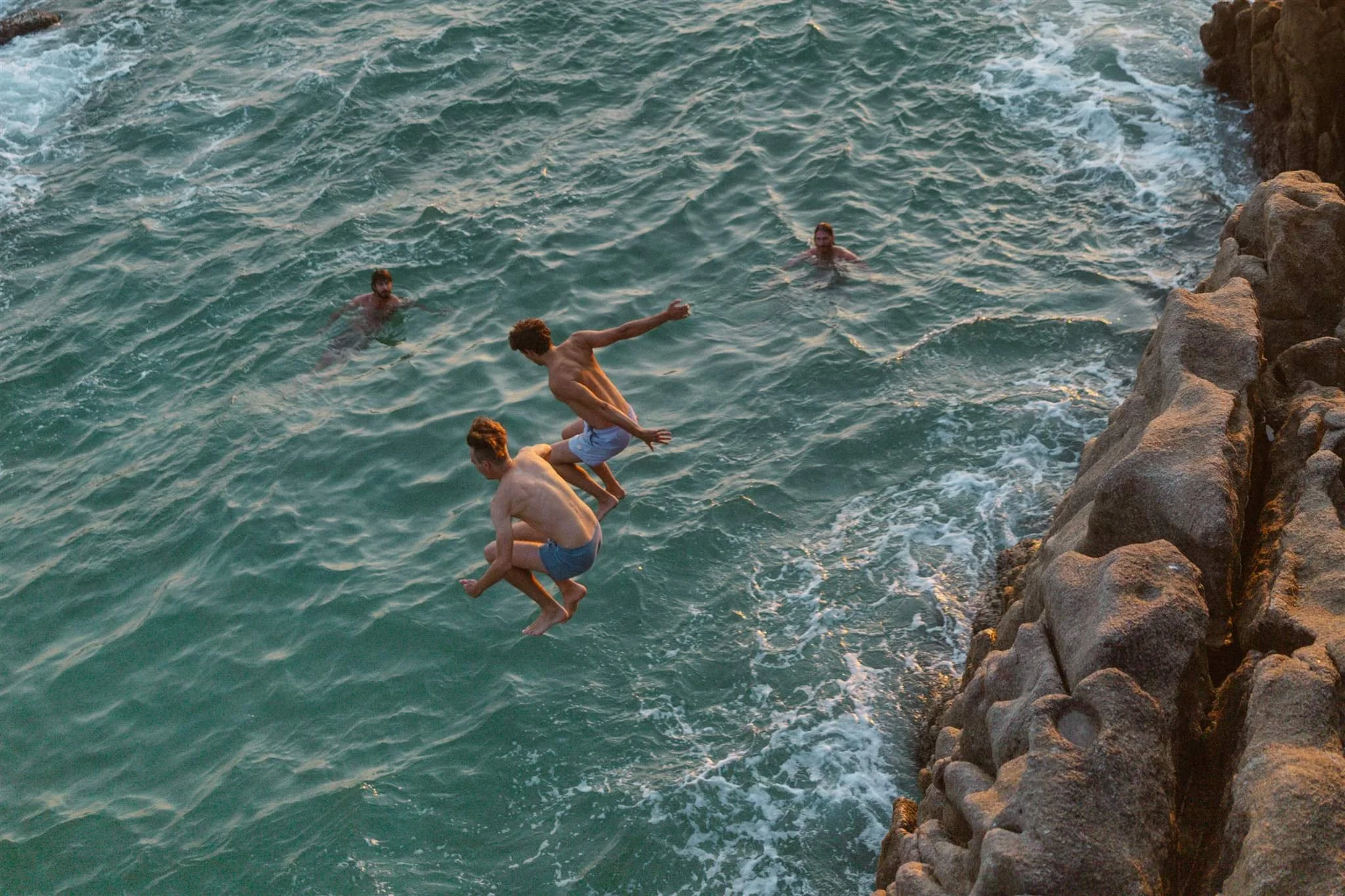 Four young men are jumping into the water from a rocky shoreline, with three already swimming in the sea.