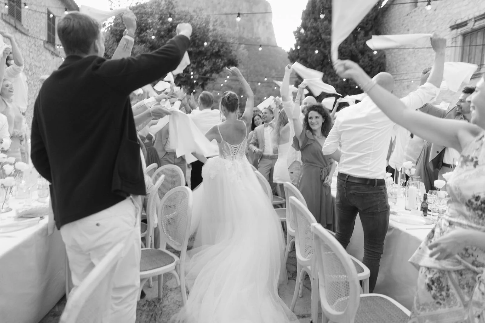 People at a wedding celebration, some waving napkins over the bride in a white wedding dress, in an outdoor venue decorated with string lights.