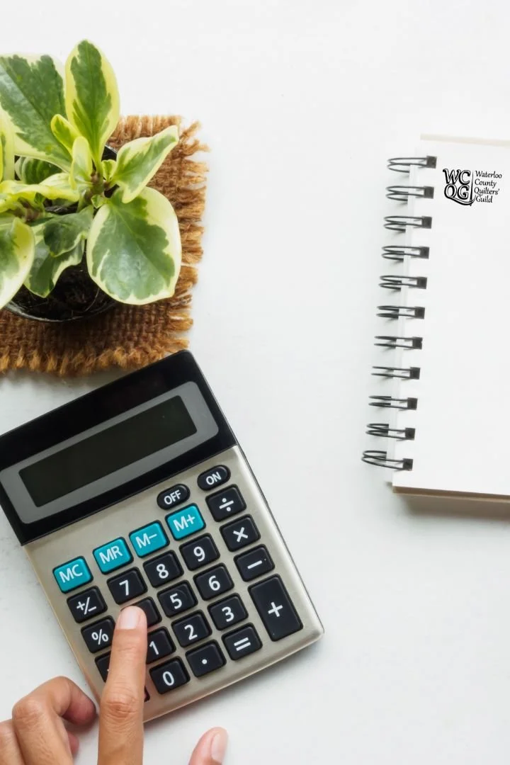 A calculator on a white surface with a person's finger pressing a button, a notebook with the Waterloo County Quilters' Guild logo, a potted plant on a woven mat.