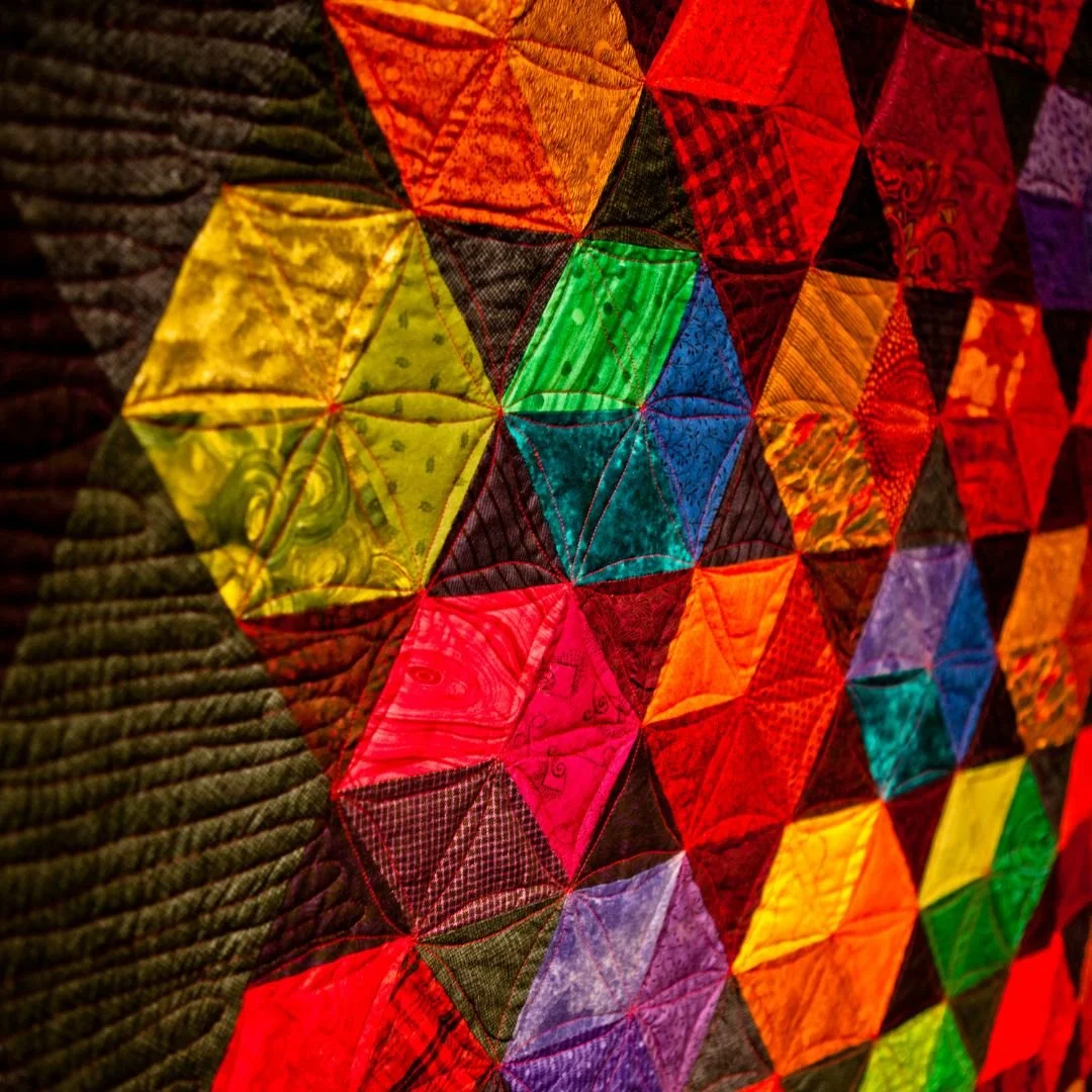 Close-up of a colorful quilt with a pattern of various vibrant squares and triangles, featuring red, yellow, green, blue, purple, and black fabric pieces.