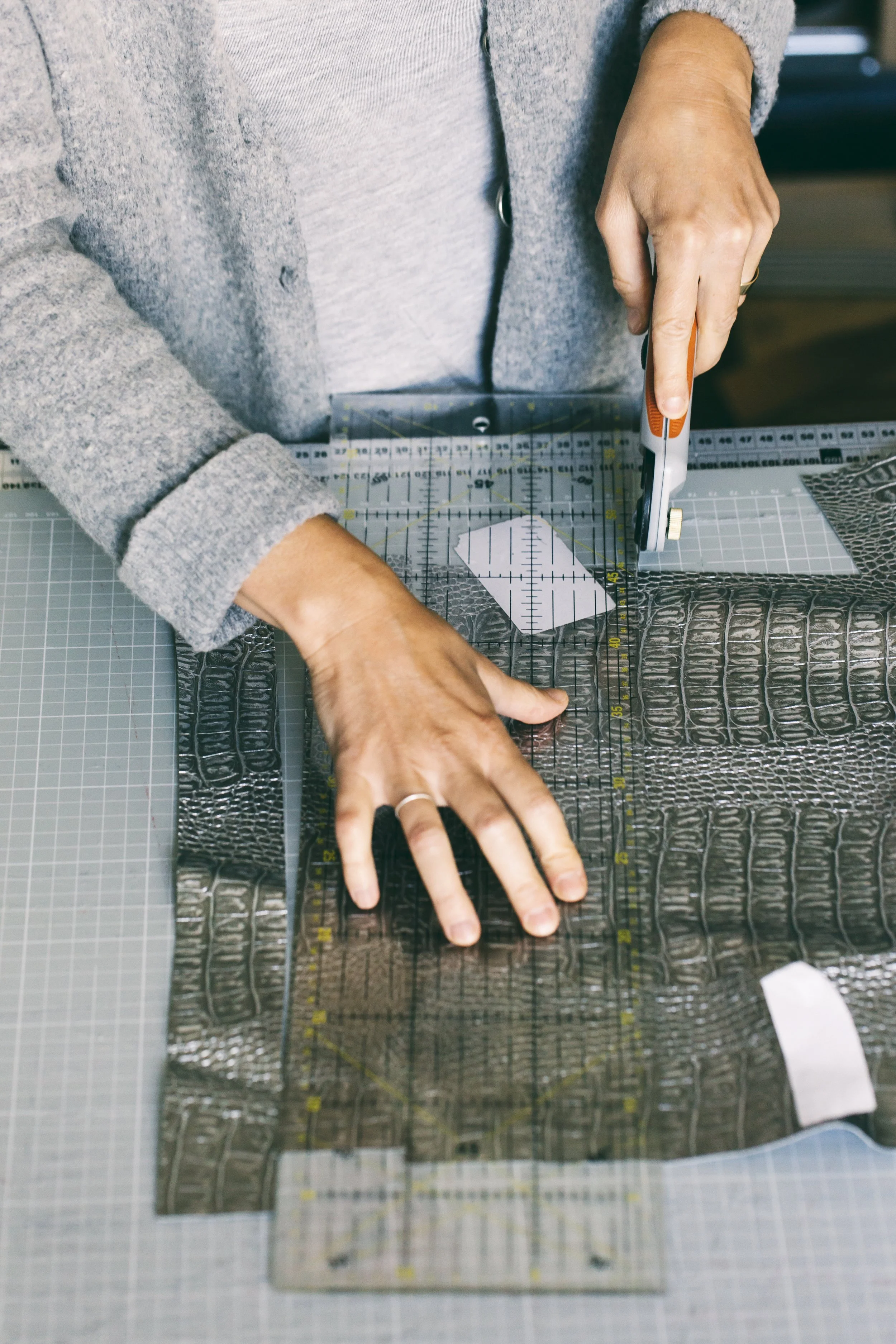 Person cutting patterned leather or faux crocodile-textured material with a rotary cutter on a cutting mat, using a clear ruler for measurements.