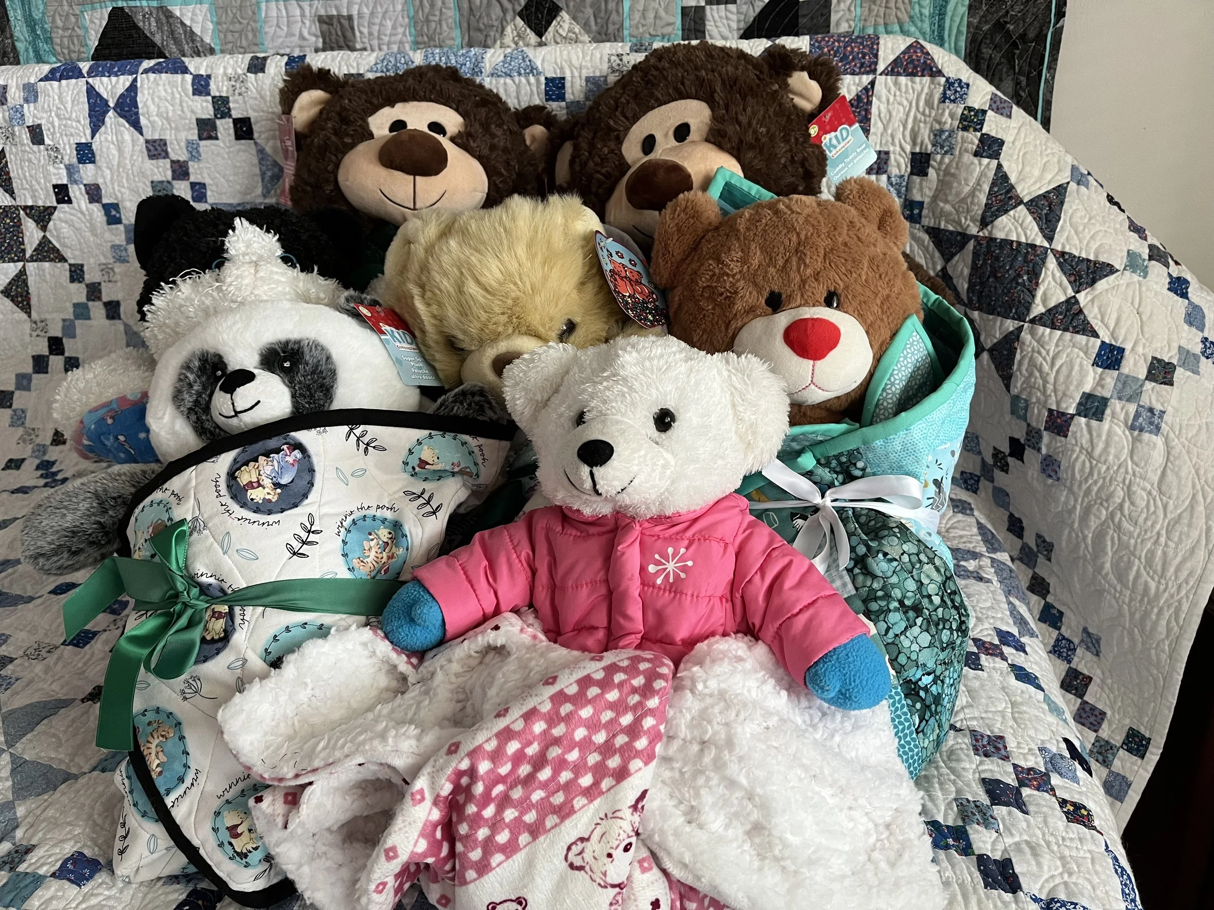 Collection of plush animal toys, including bears, a panda, and a lion, arranged on a quilted blanket with a patchwork design. One bear is dressed in a pink jacket, and some toys are in a small, decorated basket.