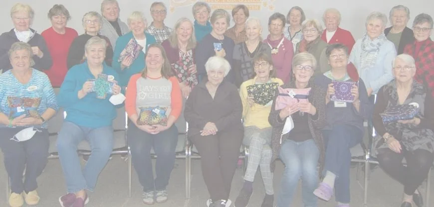 Group of women, mostly middle-aged to elderly, gathered together indoors, some holding wrapped gifts, smiling for a photo.