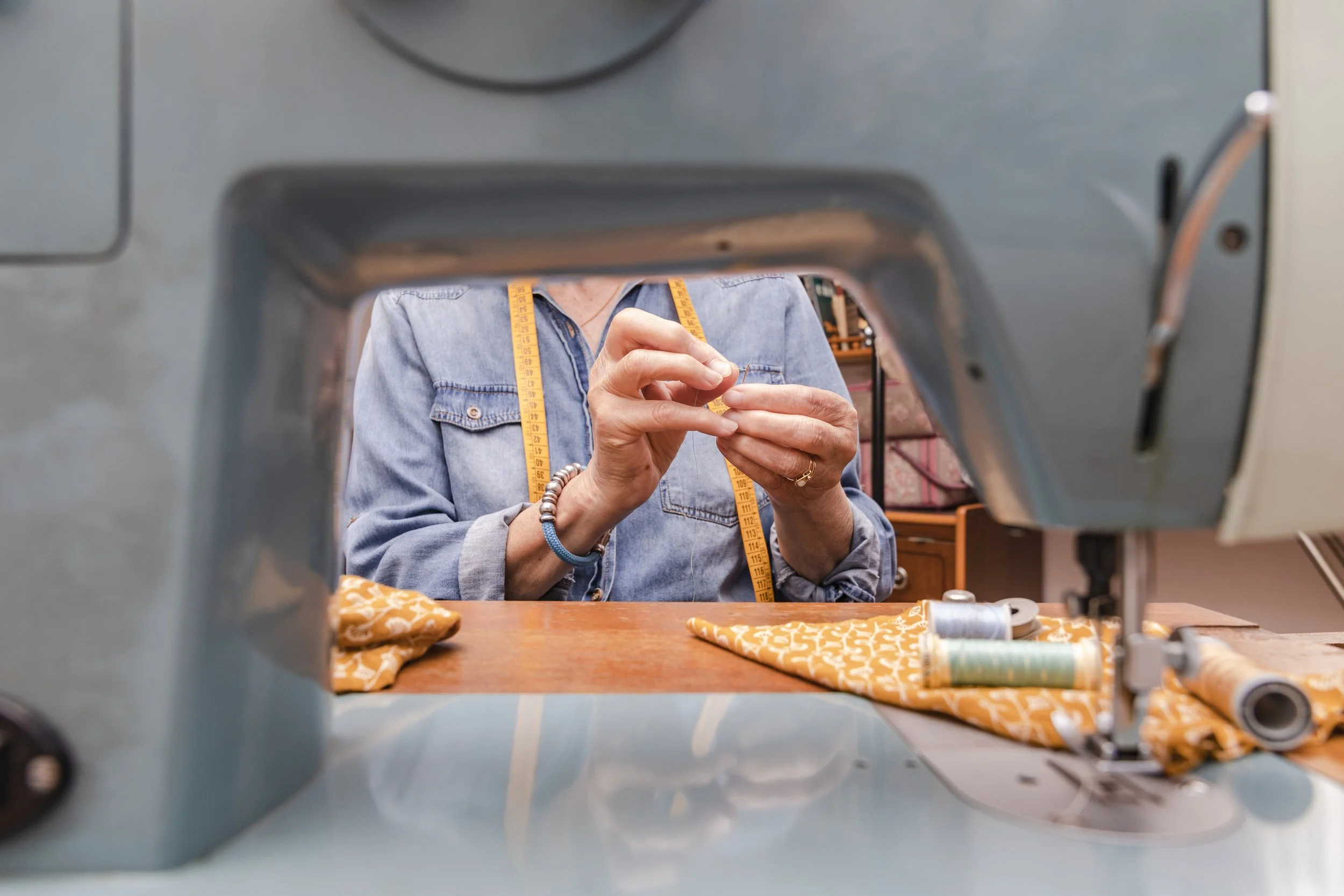 A person sewing at a sewing machine, seen from the machine's perspective, with spools of thread and fabric on the table.
