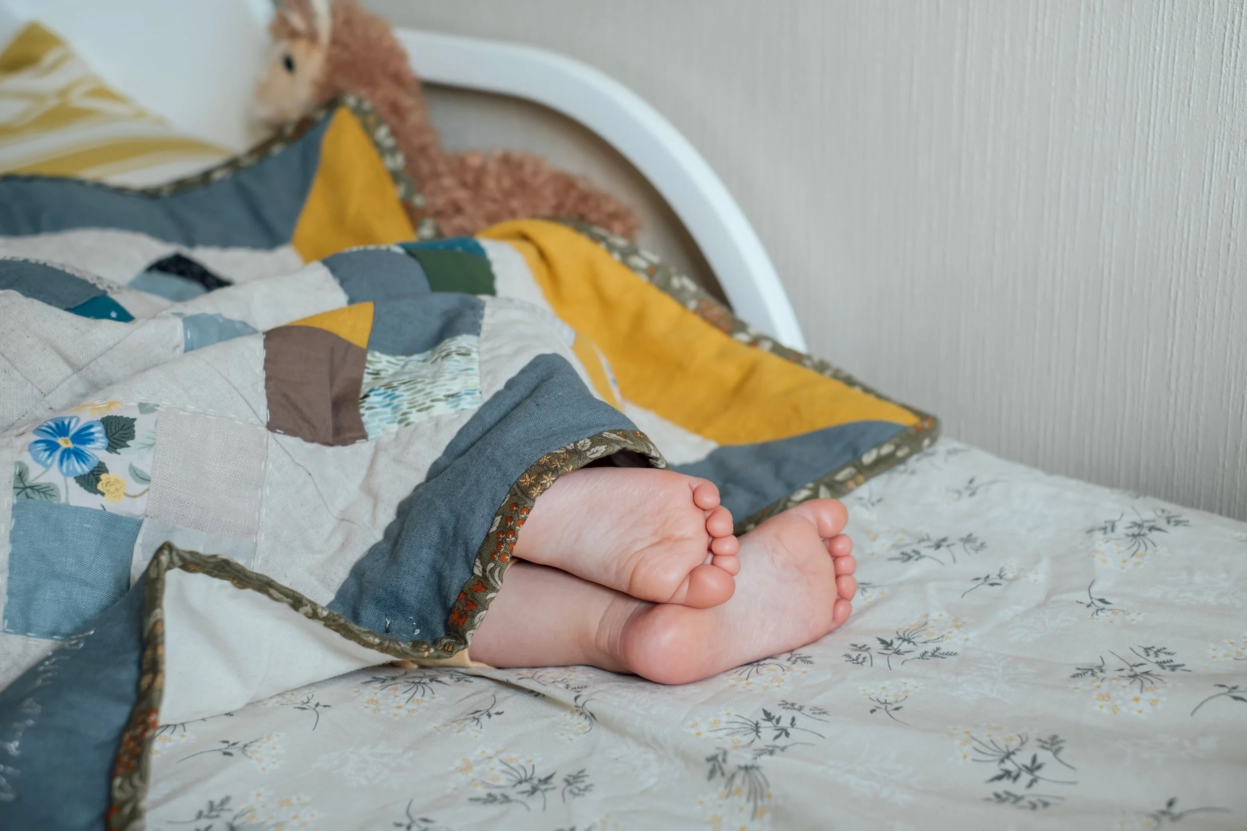 A child or baby lying in bed with only their feet visible, covered by a colorful quilt, with a teddy bear resting against the headboard.
