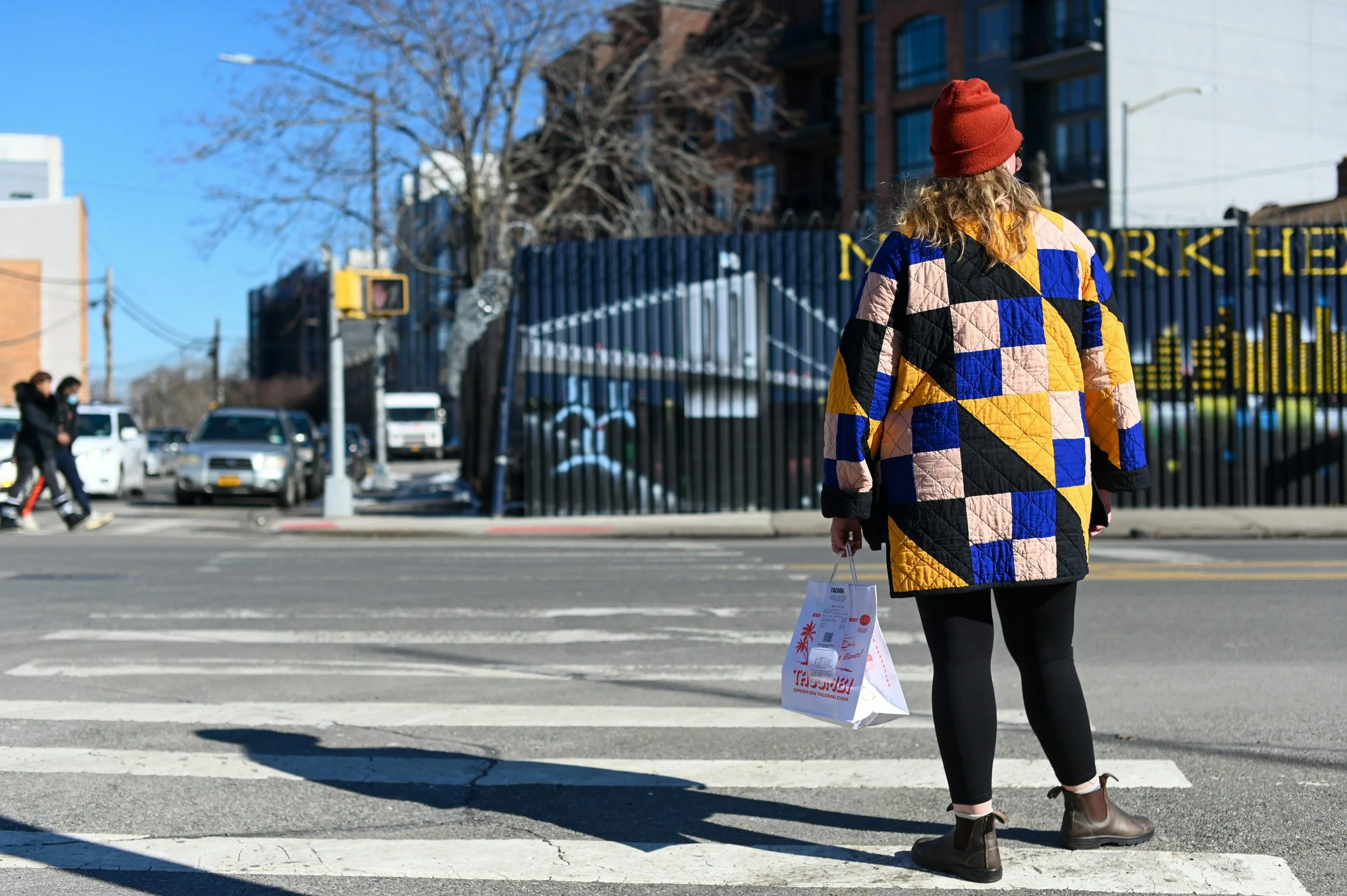 A woman in a colorful patchwork jacket and red beanie crossing the street at a crosswalk, holding a shopping bag, with cars and pedestrians in the background under a blue sky.