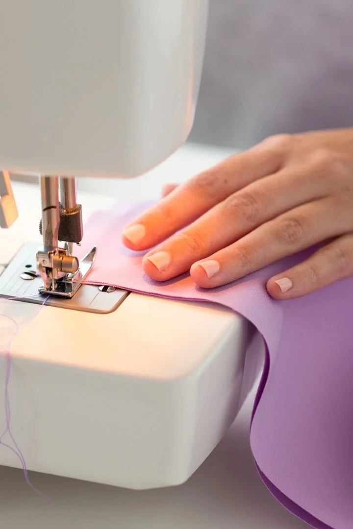 A close-up view of a person's hand guiding purple fabric through a sewing machine.