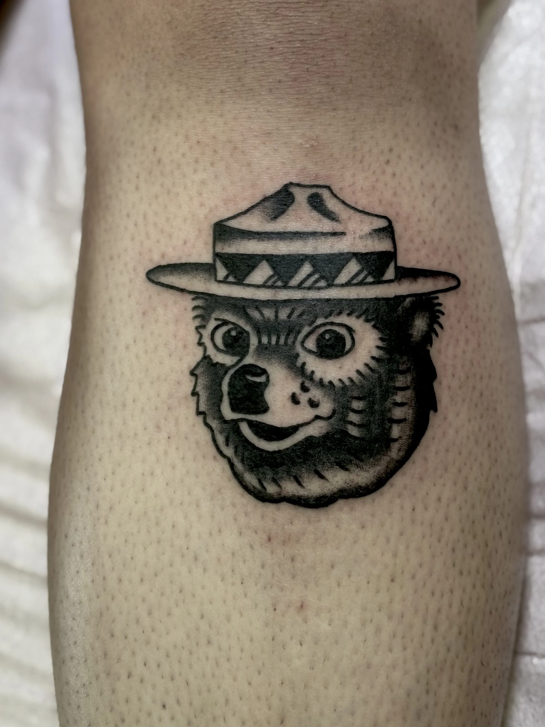 Tattoo of a Smokey the bear wearing a wide-brimmed hat with a band, on a person's skin.