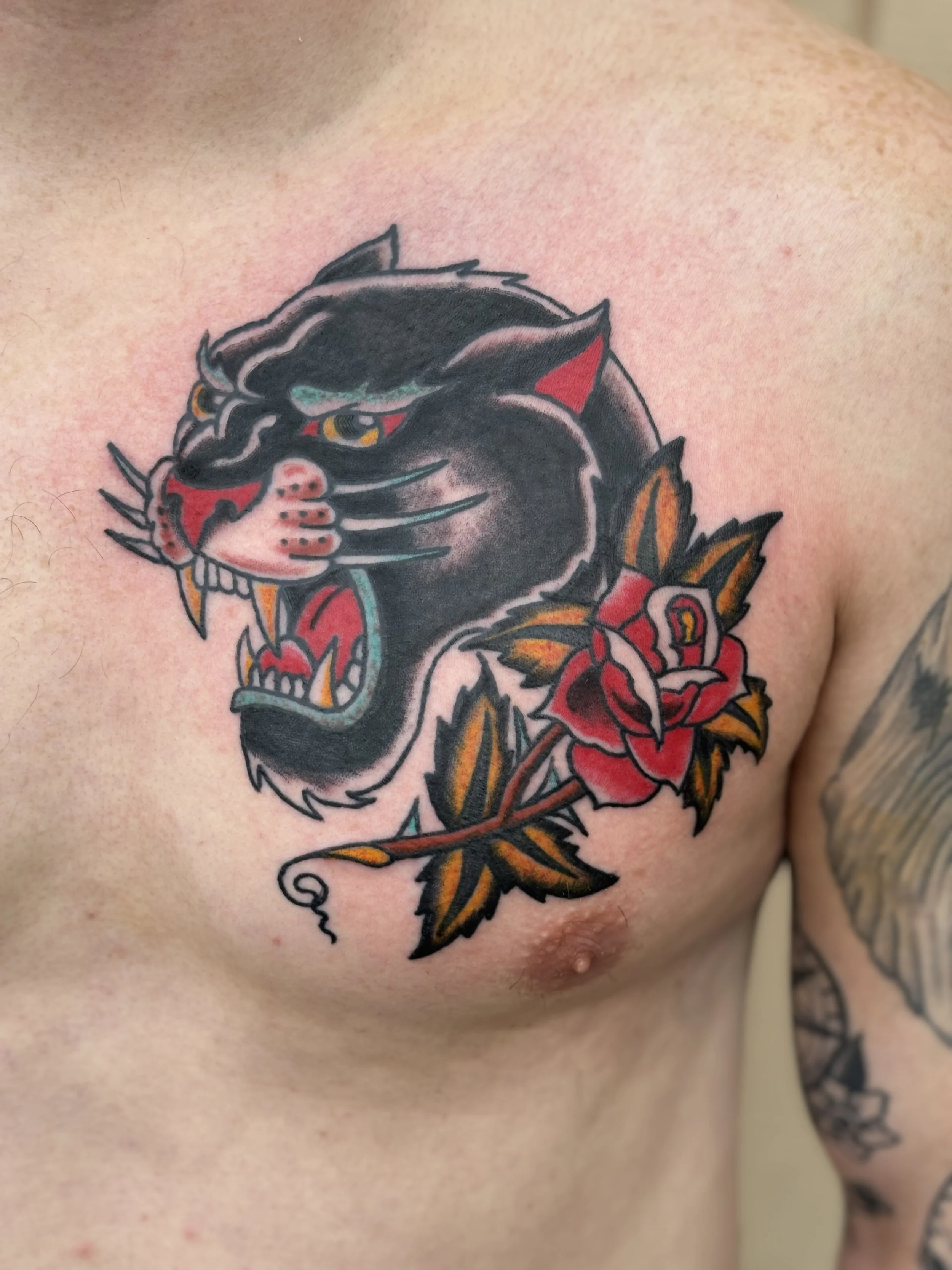 Close-up of a tattoo on a person's upper chest and shoulder area. The tattoo features a black panther's head with fierce yellow eyes, red accents on the face, and surrounding red and orange roses with green leaves.