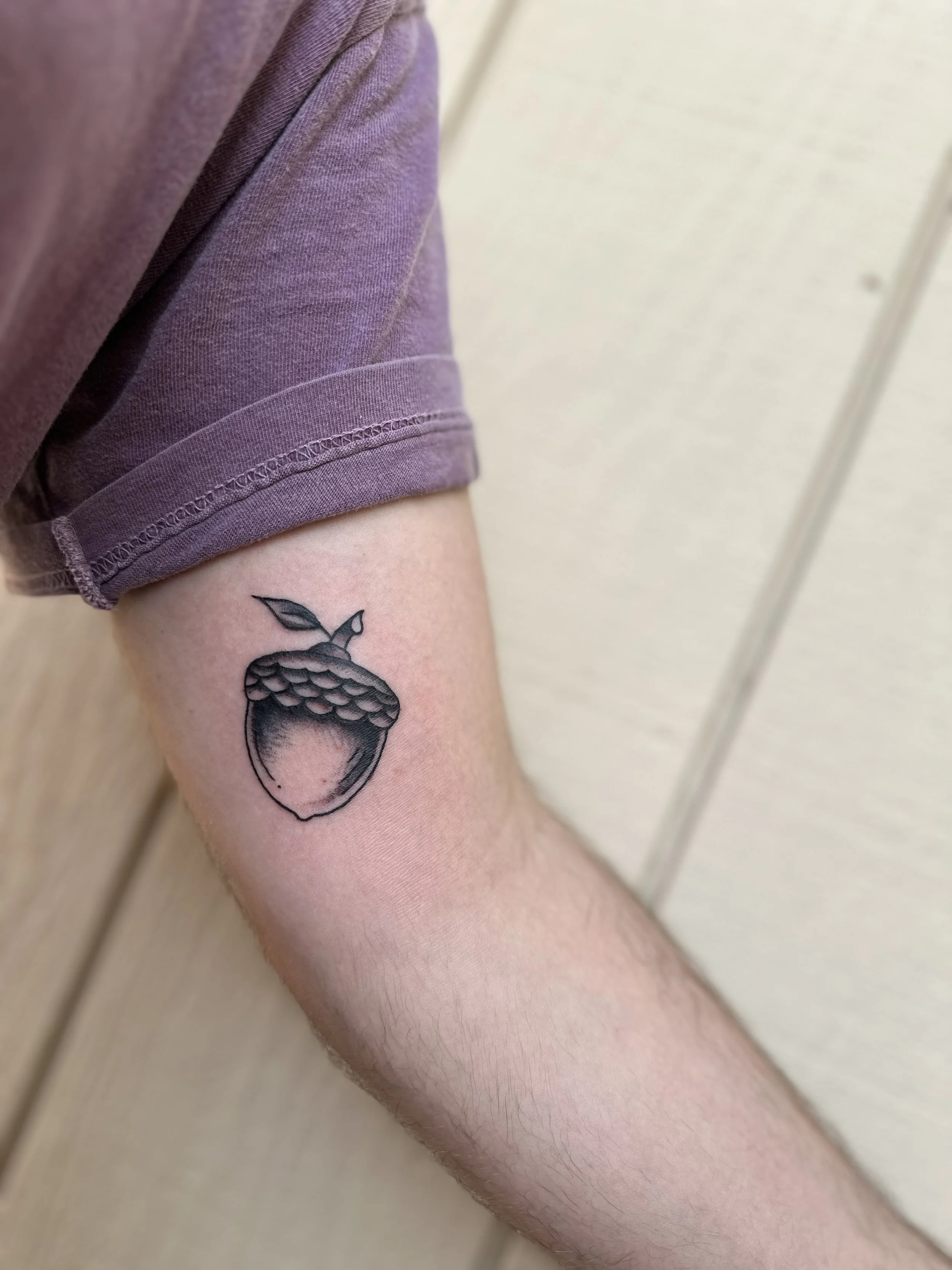 A person's arm with a tattoo of a acorn on the forearm, wearing a purple short-sleeve shirt, against a light-colored wall background.
