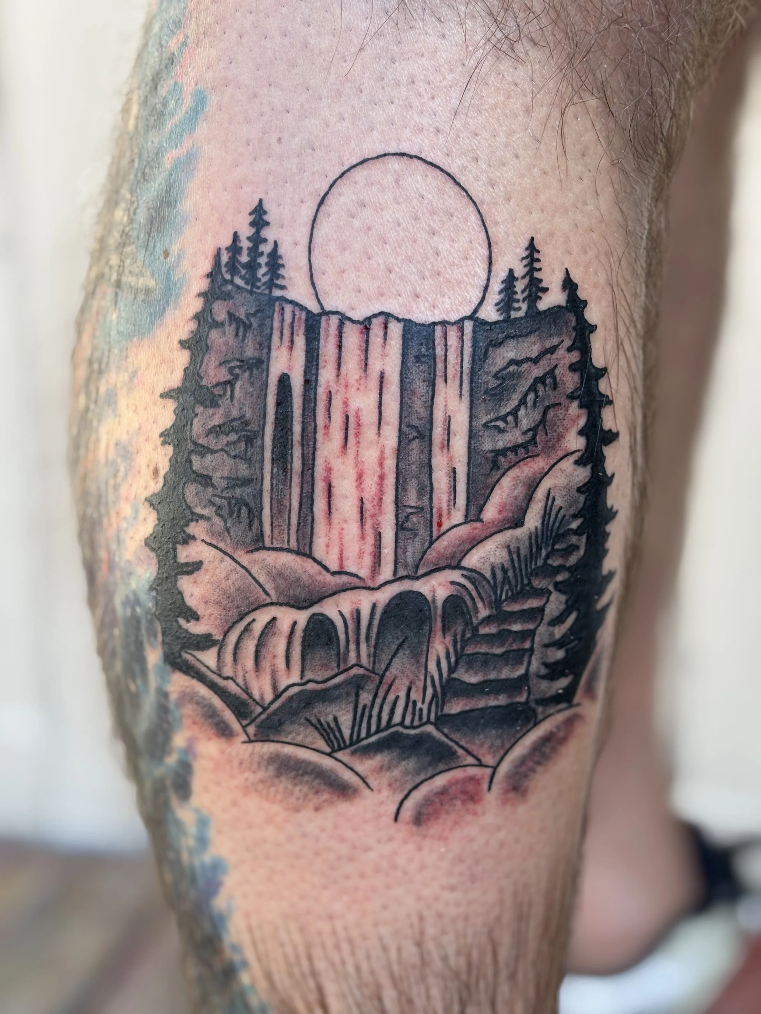 Tattoo of Vernal falls in Yosemite National Park with a waterfall and pine trees and a circular sun in the background on the person's skin.