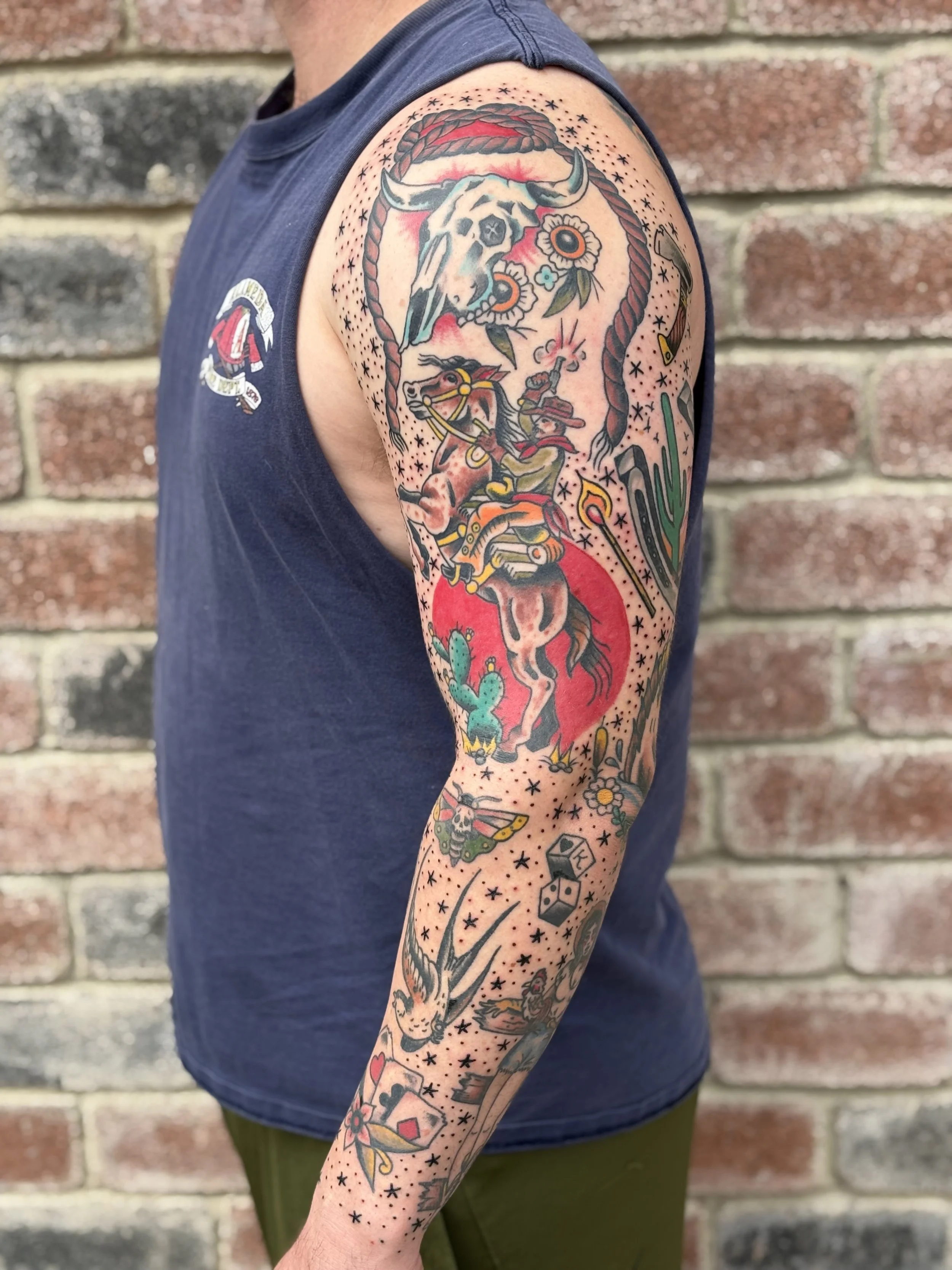 Tattoo sleeve on person's arm featuring a cow skull, flowers, a cowboy riding a bucking horse, cacti, dice, and other motifs in traditional tattoo style.