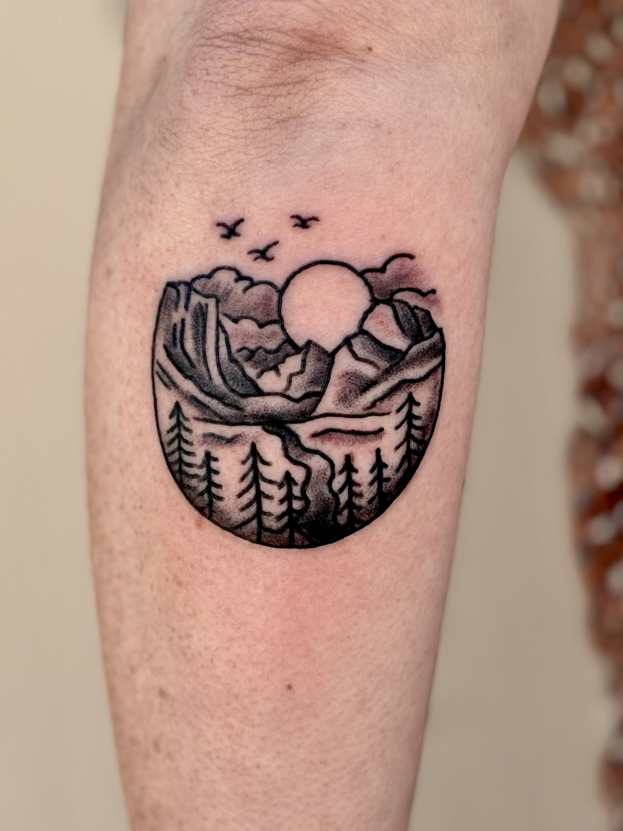 Tattoo of a Inspiration point in Yosemite with trees, clouds, and a sun, on someone's arm.