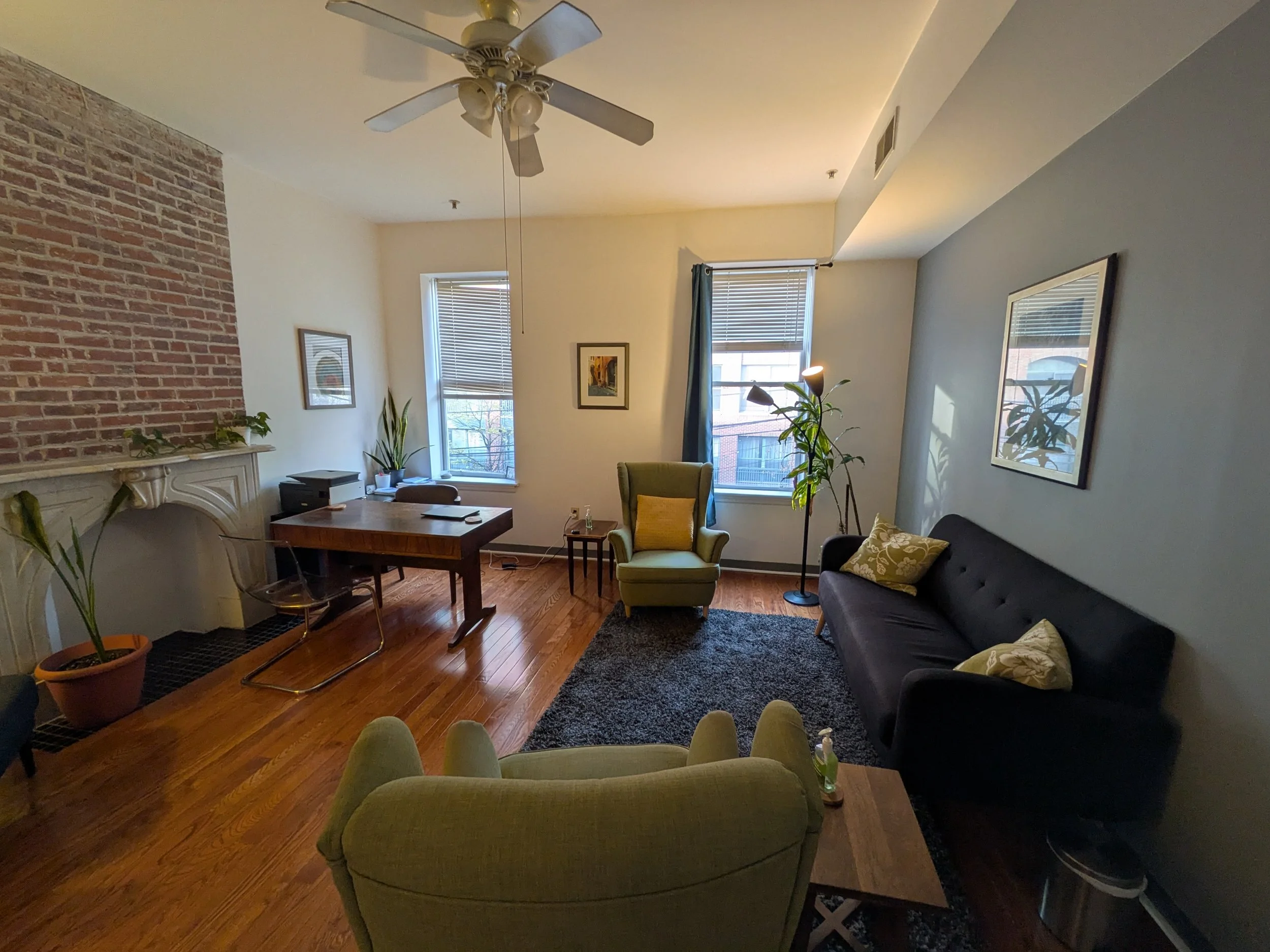 Living room with wooden floor, a black couch with yellow and patterned cushions, a green armchair with a yellow cushion, a tall lamp, artwork on the walls, two windows with blinds, and a ceiling fan.