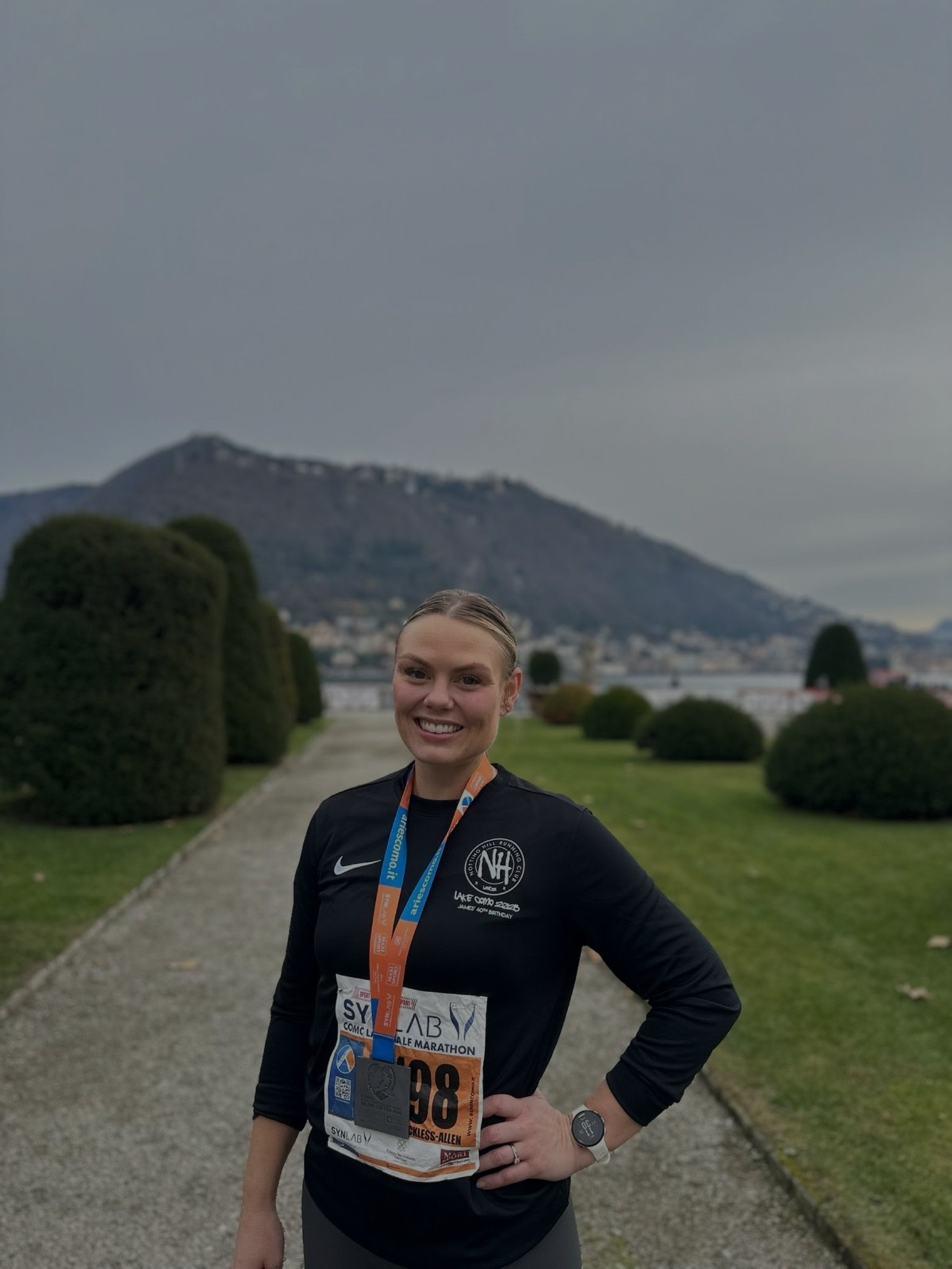 A smiling female runner in black athletic attire with a race bib and medal stands outdoors on a path with trimmed bushes and trees, with a mountain and cloudy sky in the background.