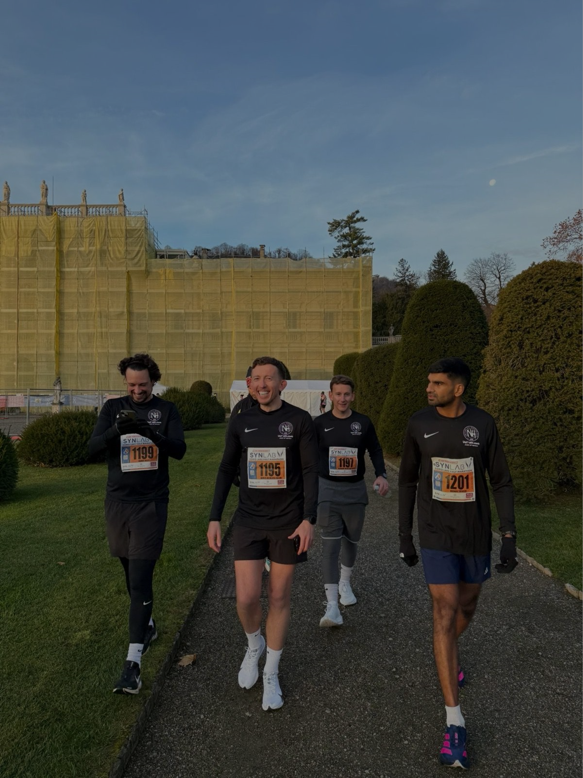 Four runners wearing black athletic gear jogging on a path during a race, with a large building covered in scaffolding and yellow netting behind them, and neatly trimmed bushes on either side.