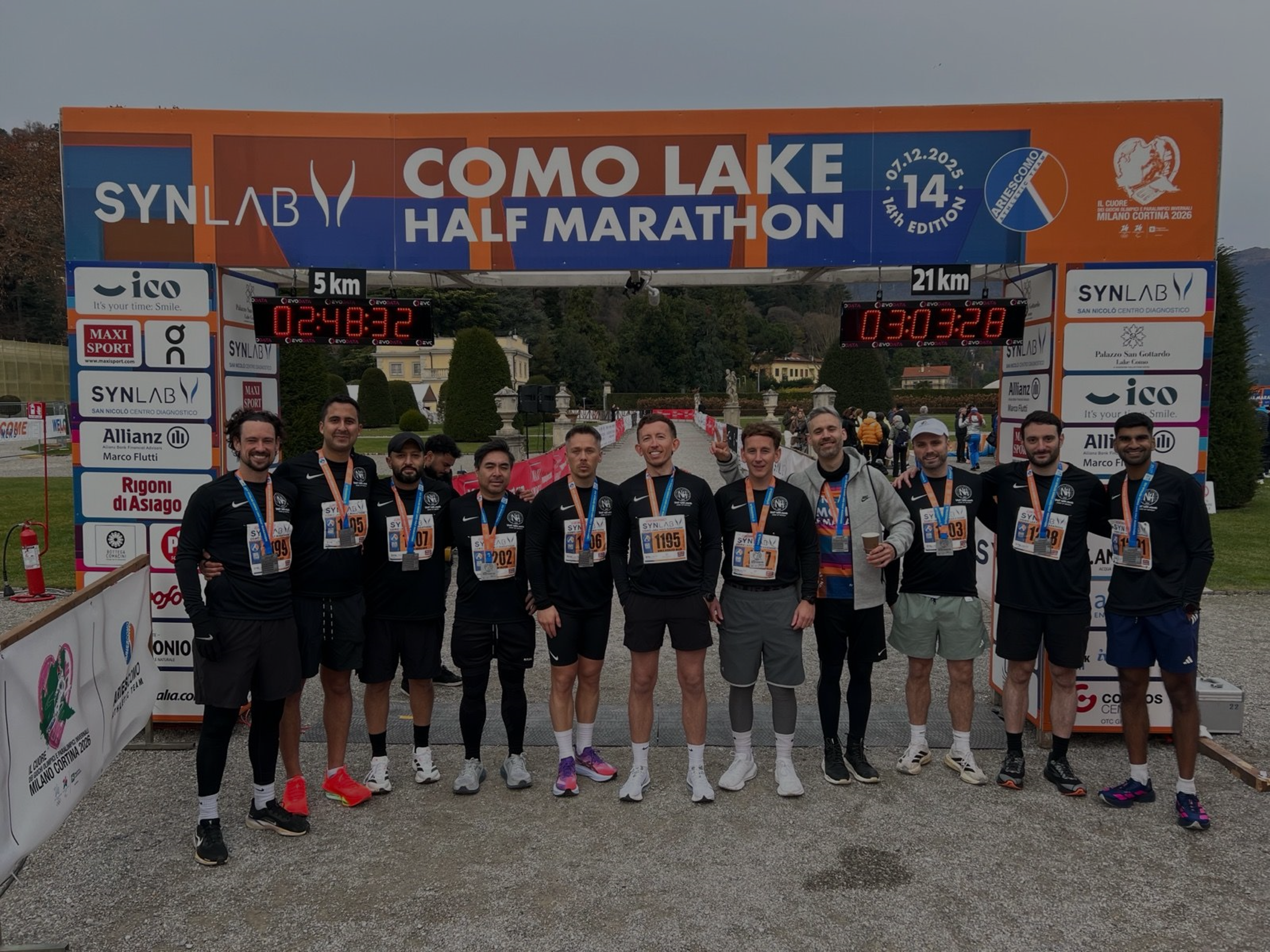 Group of runners posing with medals after completing the Como Lake Half Marathon, standing under a race banner with timing clocks showing 5 km and 21 km split times.